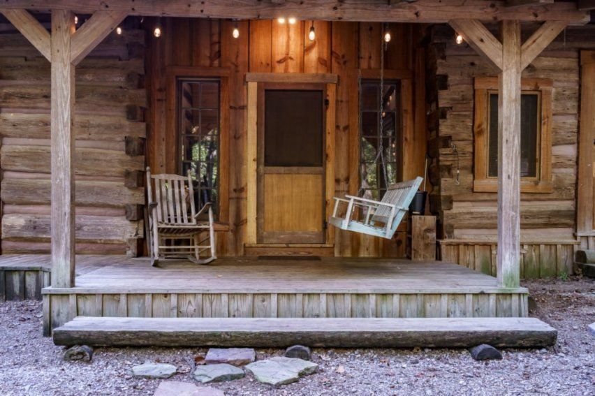 A wooden rustic cabin porch featuring a rocking chair, a hanging swing, and string lights above the doorway.