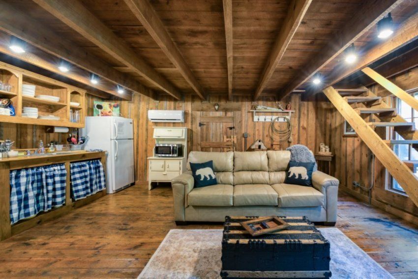 A rustic living room with exposed wood walls and beams, a beige couch with bear pillows, a kitchen area, and stairs.