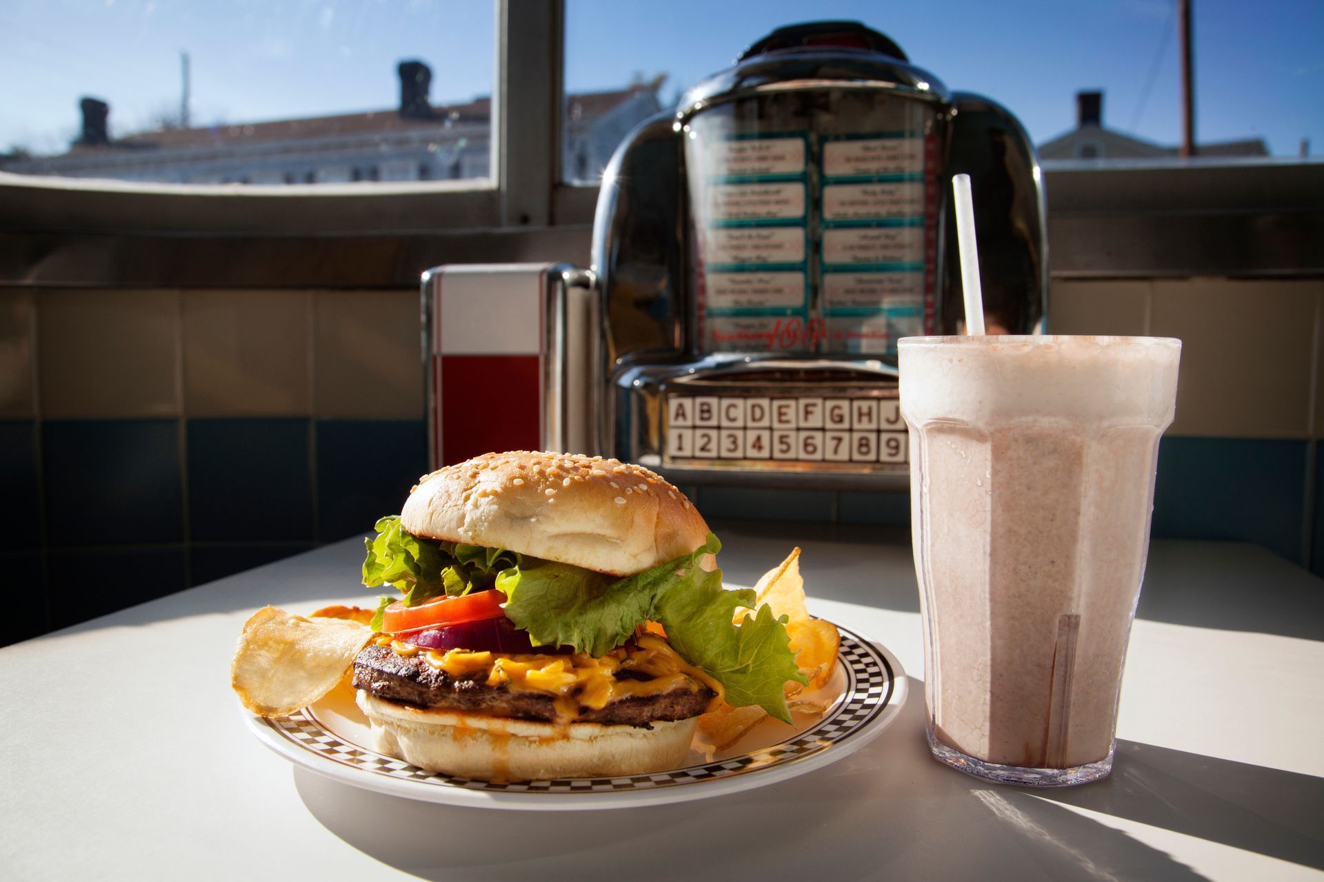 A cheeseburger with lettuce and chips on a plate, alongside a milkshake, in a retro diner with a jukebox.