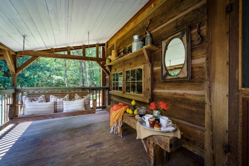 A rustic porch with a wooden swing, a bench holding drinks and flowers, and a wall-mounted mirror.