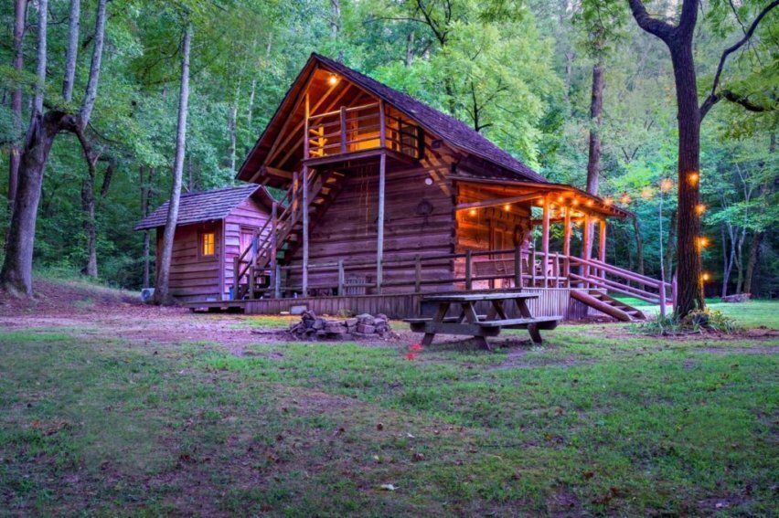 A rustic log cabin with an upper balcony and glowing string lights, nestled in a lush, green forest at dusk.