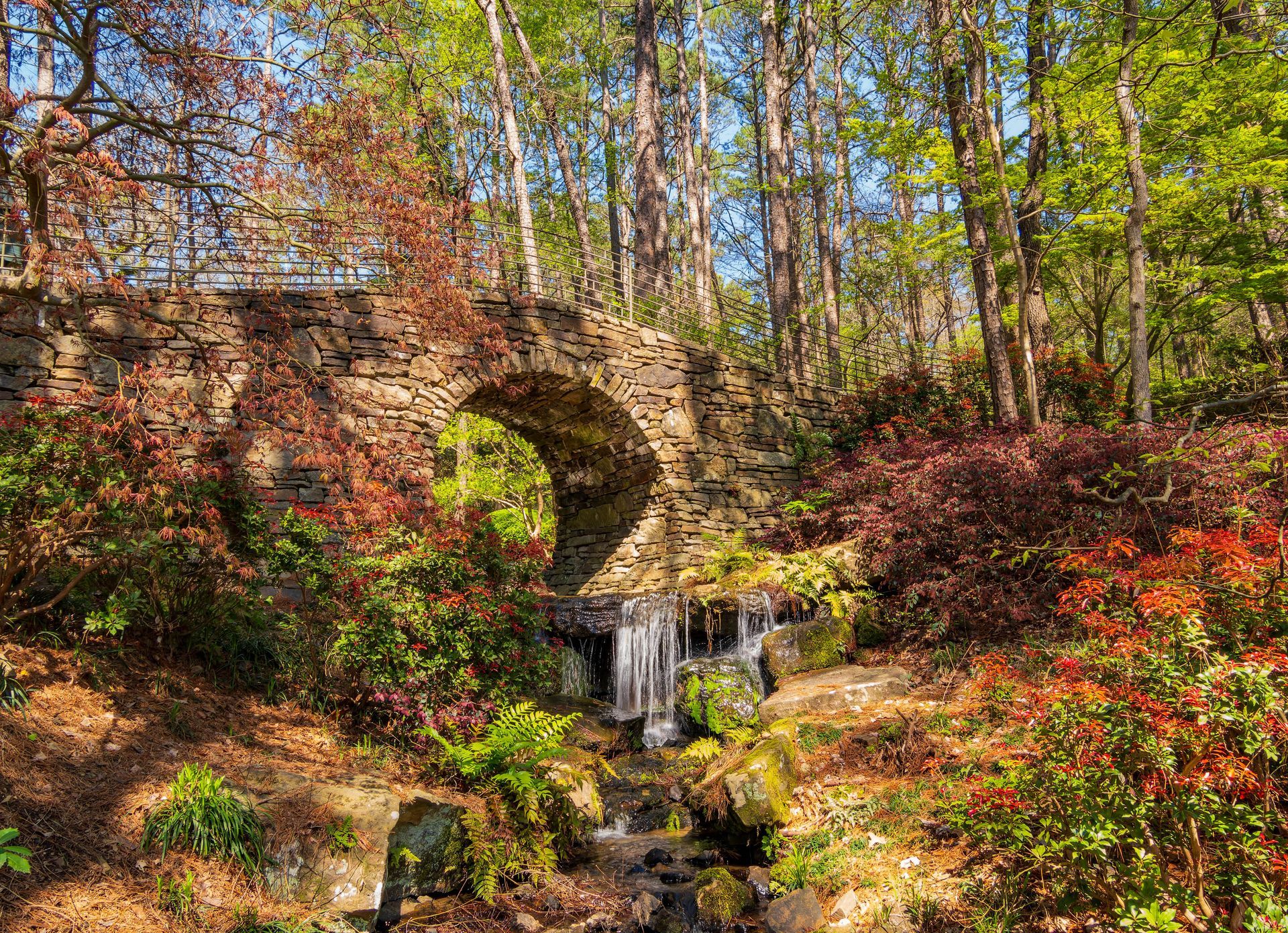 A stone arch bridge over a small waterfall in a lush forest with colorful autumn foliage.