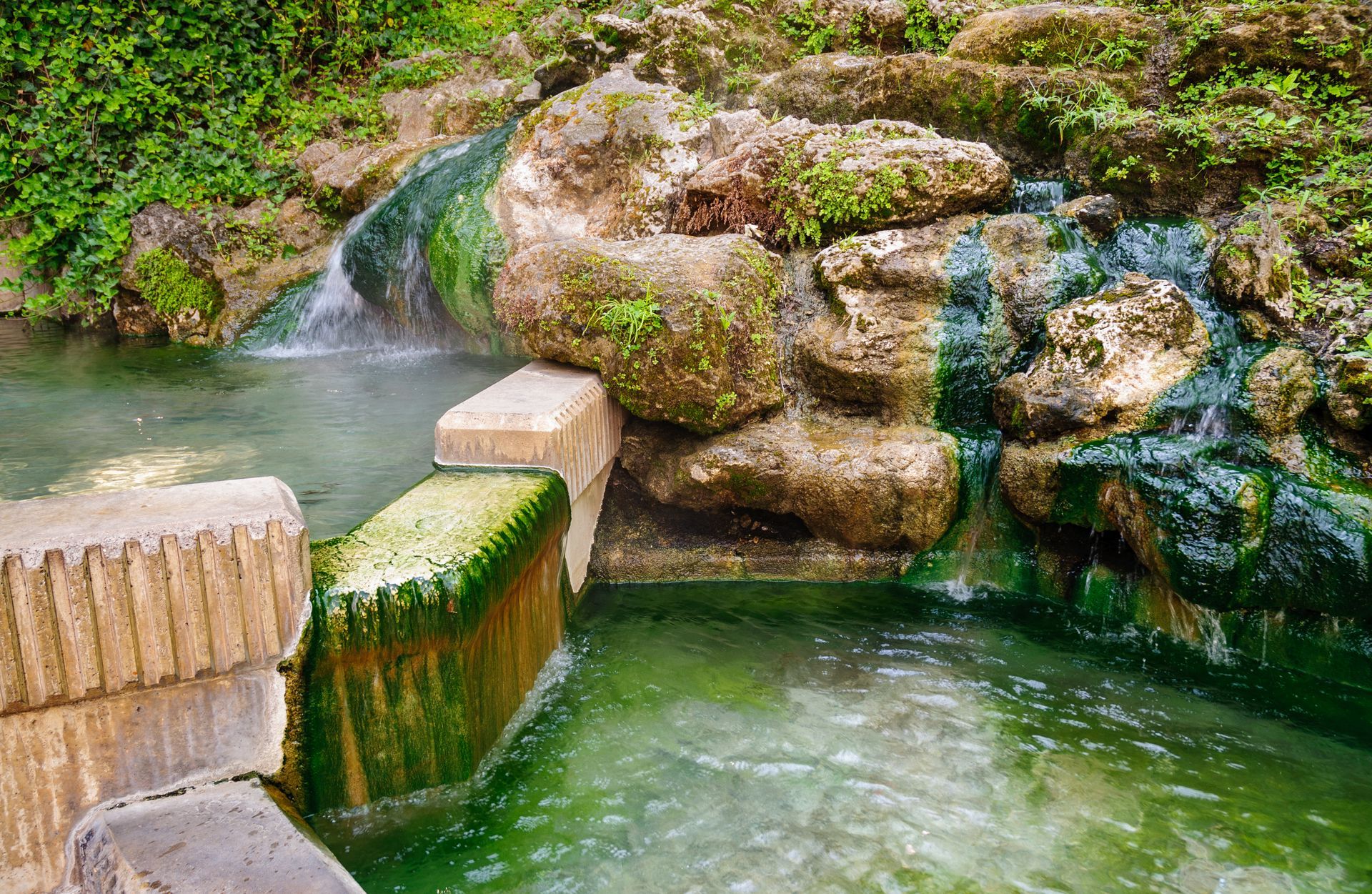 A natural spring flowing over mossy, textured rocks into a stone-lined pool, with steam rising from the water.
