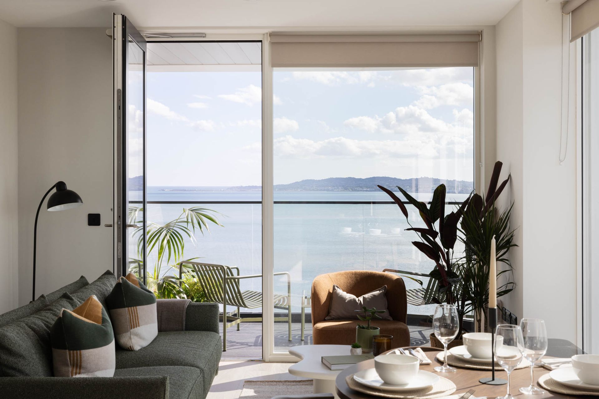Bright living room with sofa, dining table, plants, and large windows overlooking a calm ocean view
