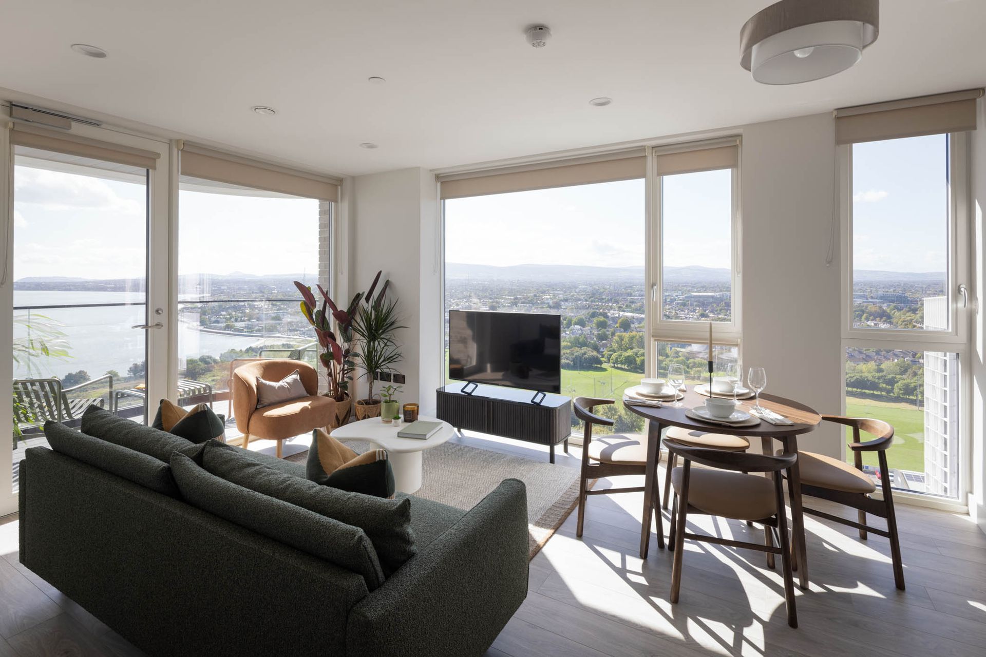 Bright living room with large windows, city view, green sofa, lounge chairs, and dining table.