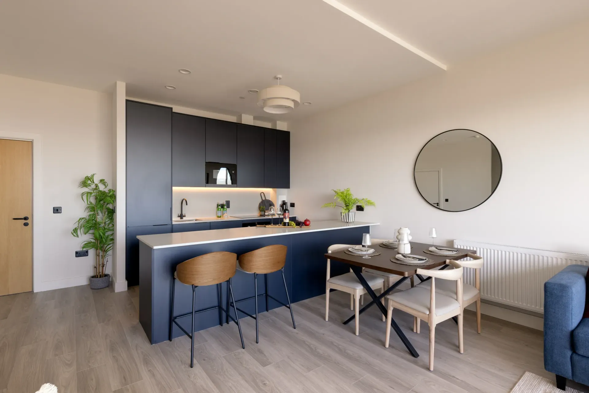 Modern open-plan kitchen and dining area with blue cabinets, wooden bar stools, round mirror, and set dining table