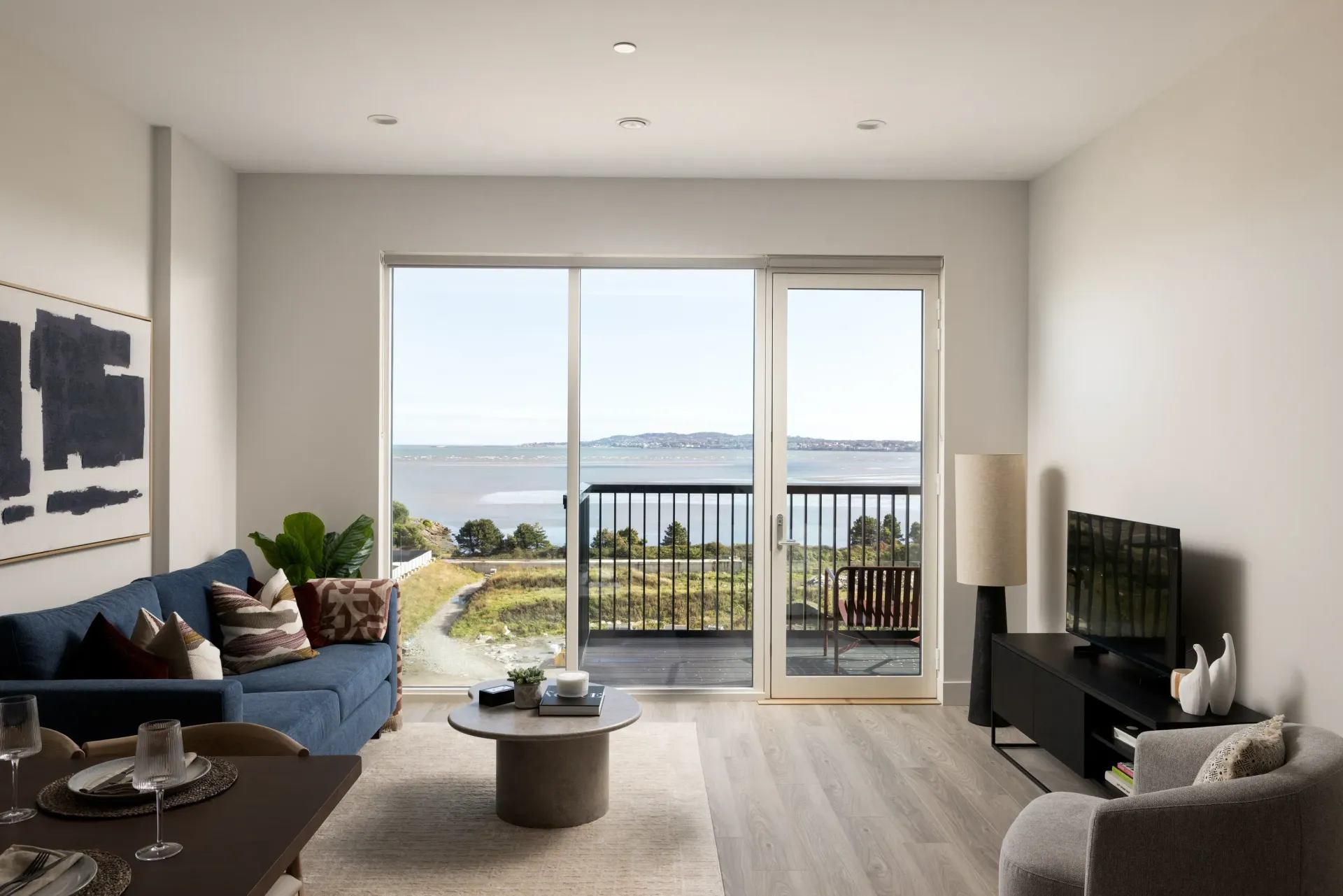 Bright living room with balcony view of water, featuring gray furniture and a TV.