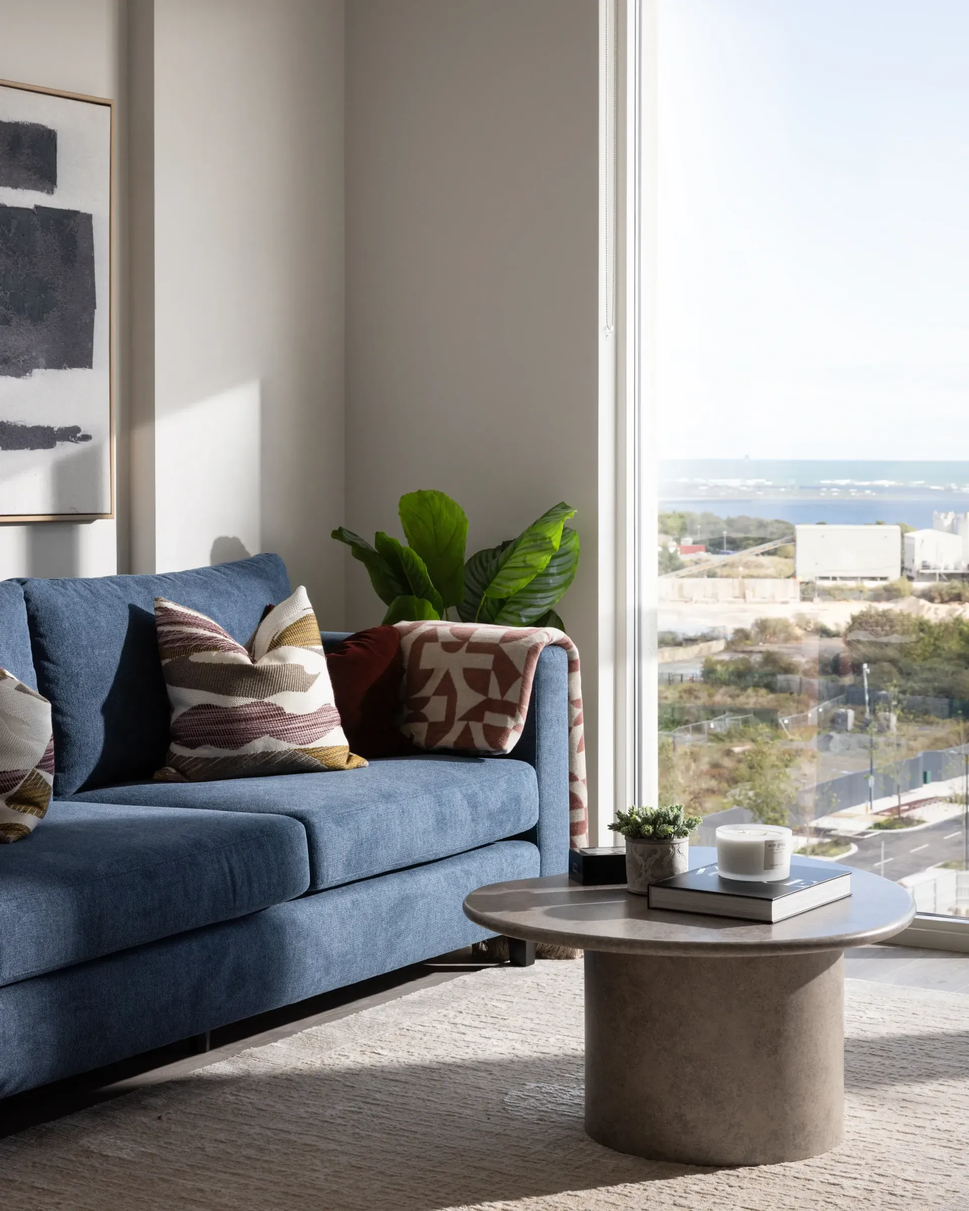 Blue sofa by a window with patterned pillows, a plant, and a small round coffee table overlooking a city view