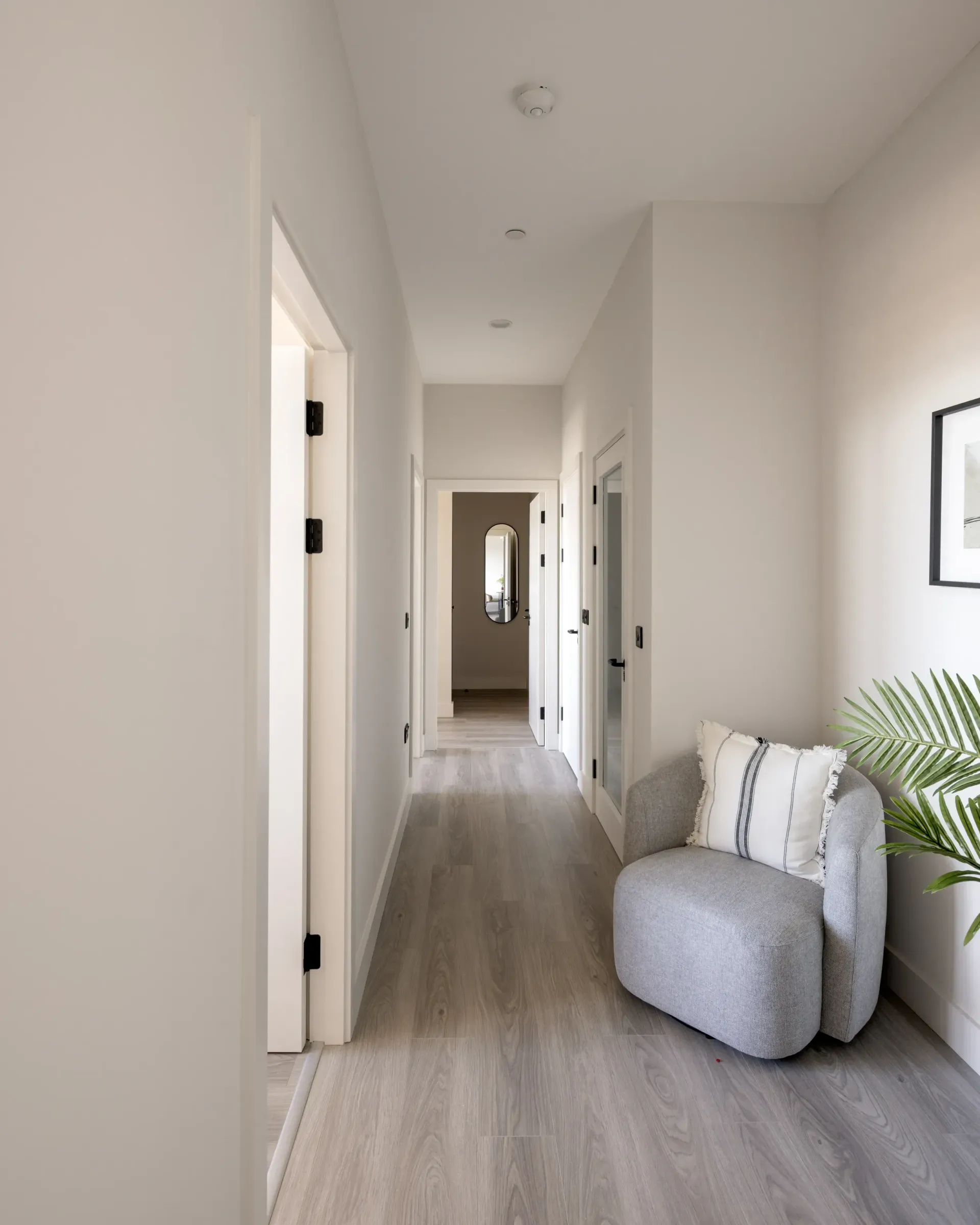 Bright hallway with light wood floors, white walls, framed art, and a gray chair by a potted plant.