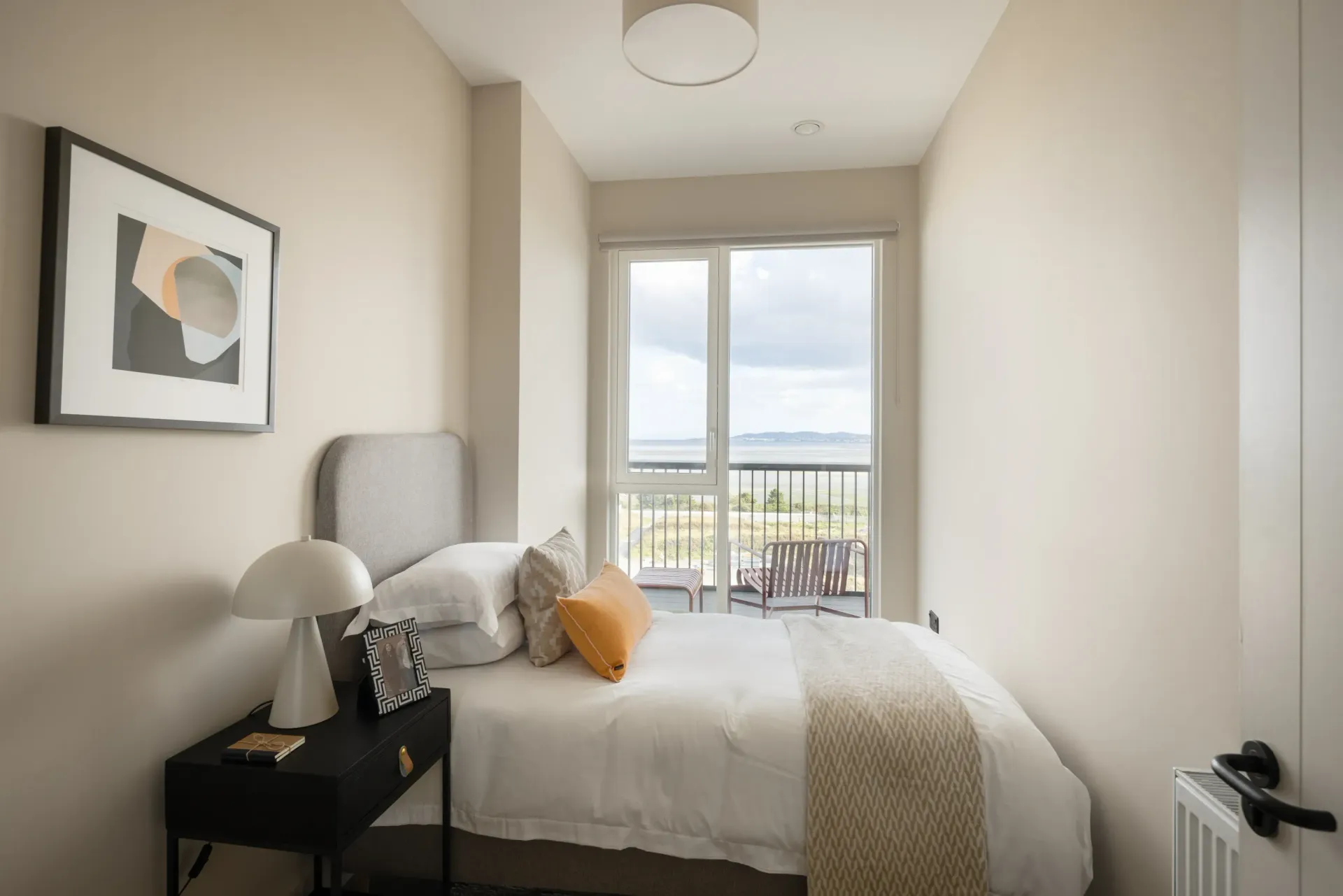 Bright minimalist bedroom with bed, side table, framed art, and window view of a balcony and sky