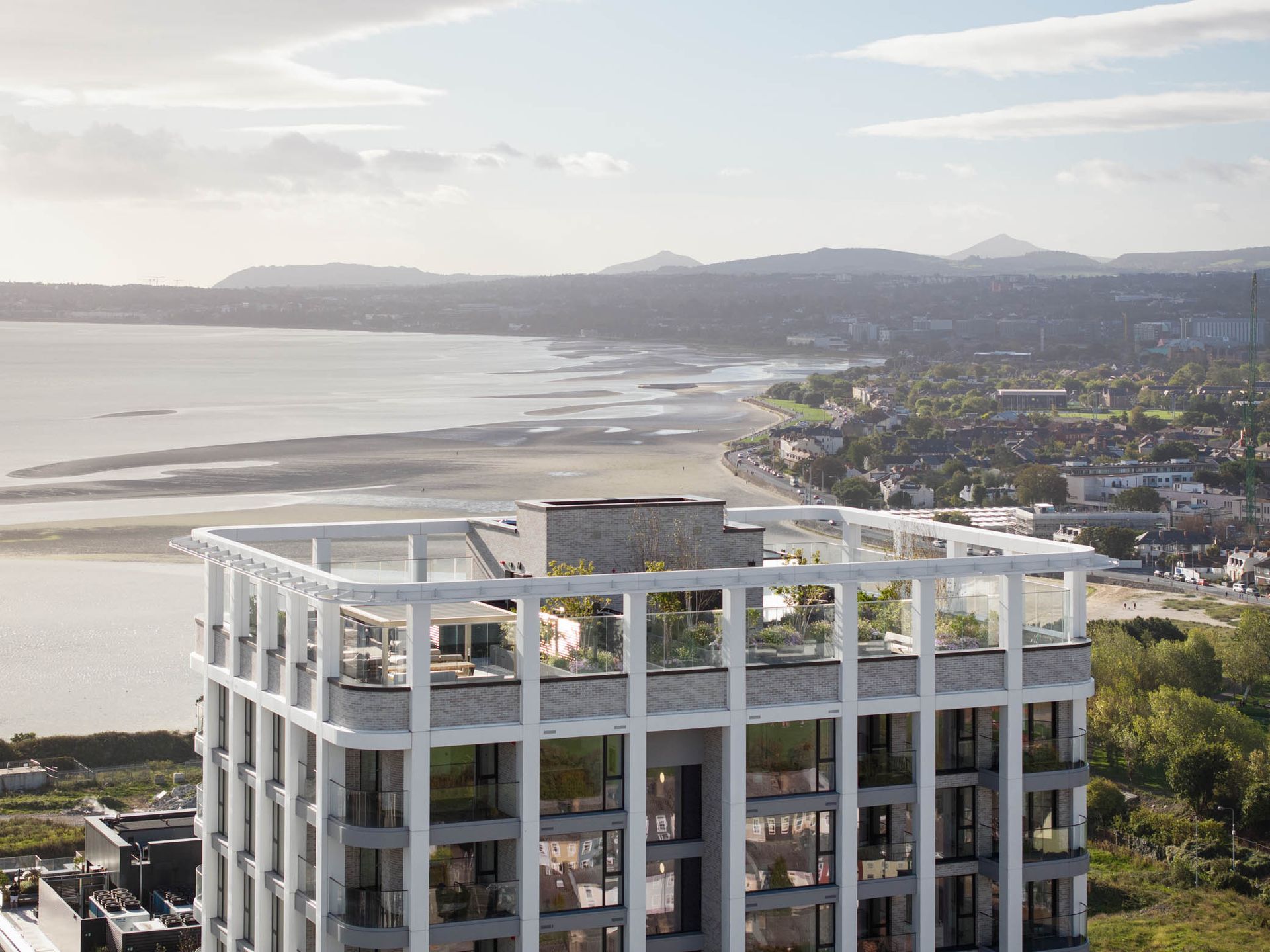 Coastal cityscape at sunset with a modern apartment building in the foreground and bay, beach, and hills beyond
