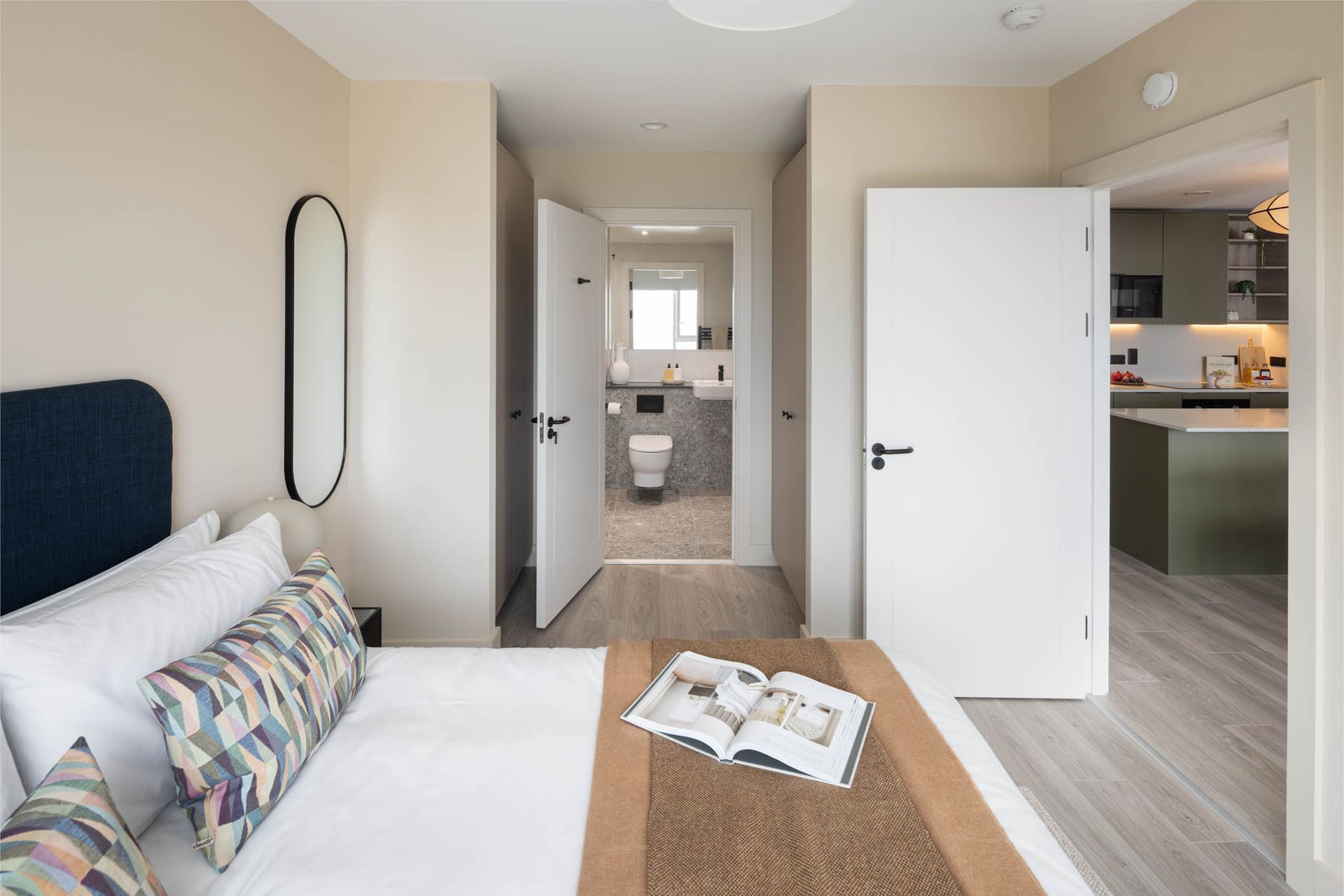 Bright bedroom with open doors to a bathroom and kitchen area, white bed, and neutral decor
