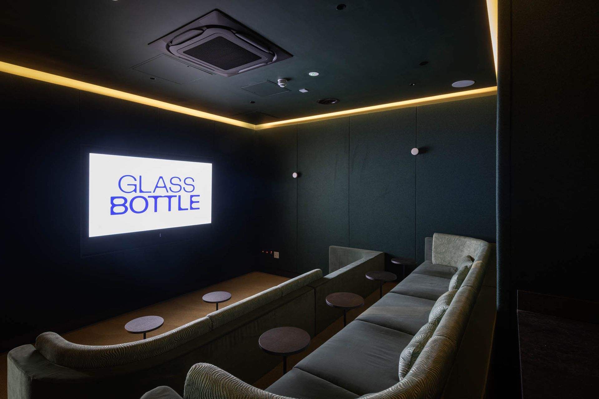 Dim lounge with dark walls, gray sofas, wood table, and a lit “GLASS BOTTLE” screen on the left.