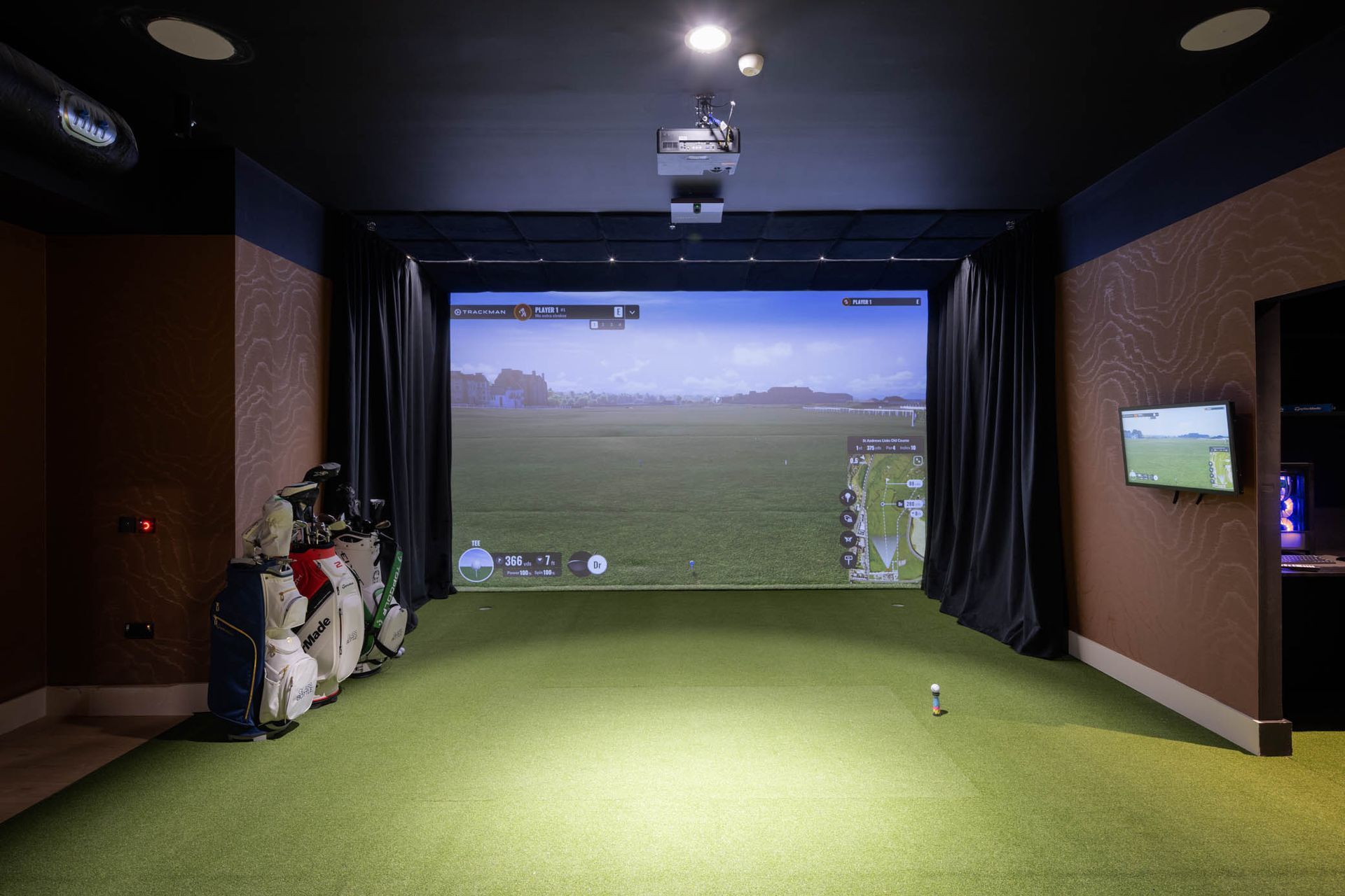 Indoor golf simulator room with green turf, large projection screen, and golf bag by the wall
