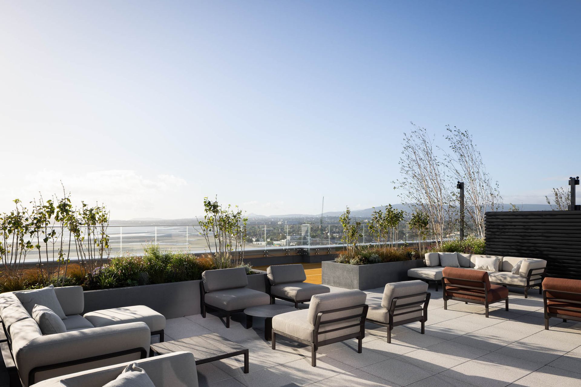 Rooftop patio with gray lounge chairs, potted plants, and a city skyline view under a clear blue sky