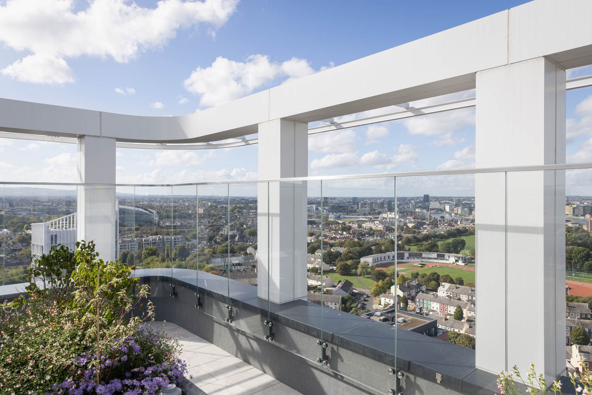 Rooftop terrace with glass railing and city skyline view under a bright blue sky.