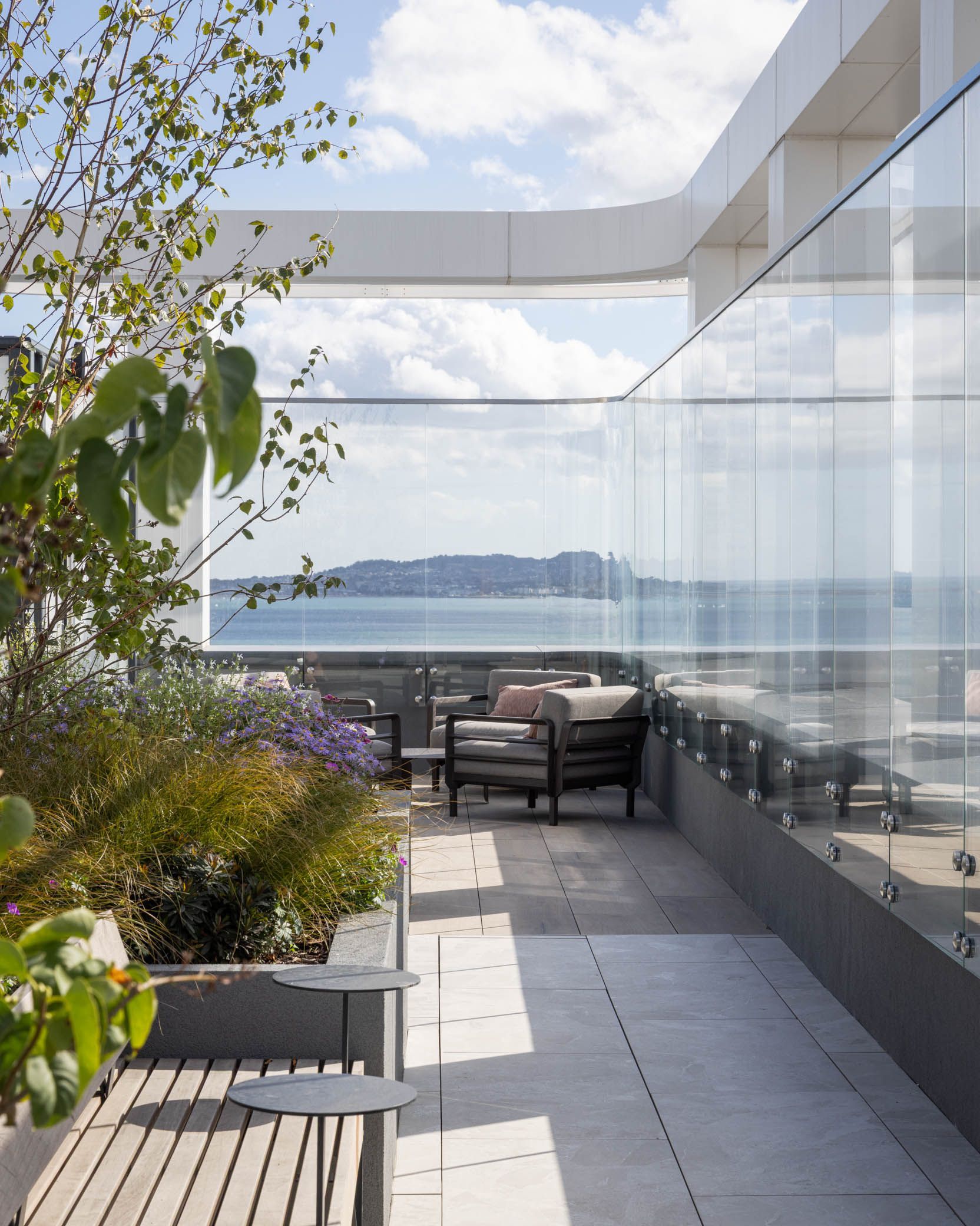 Rooftop terrace with lounge chairs, glass railing, and a view of the water under a cloudy sky