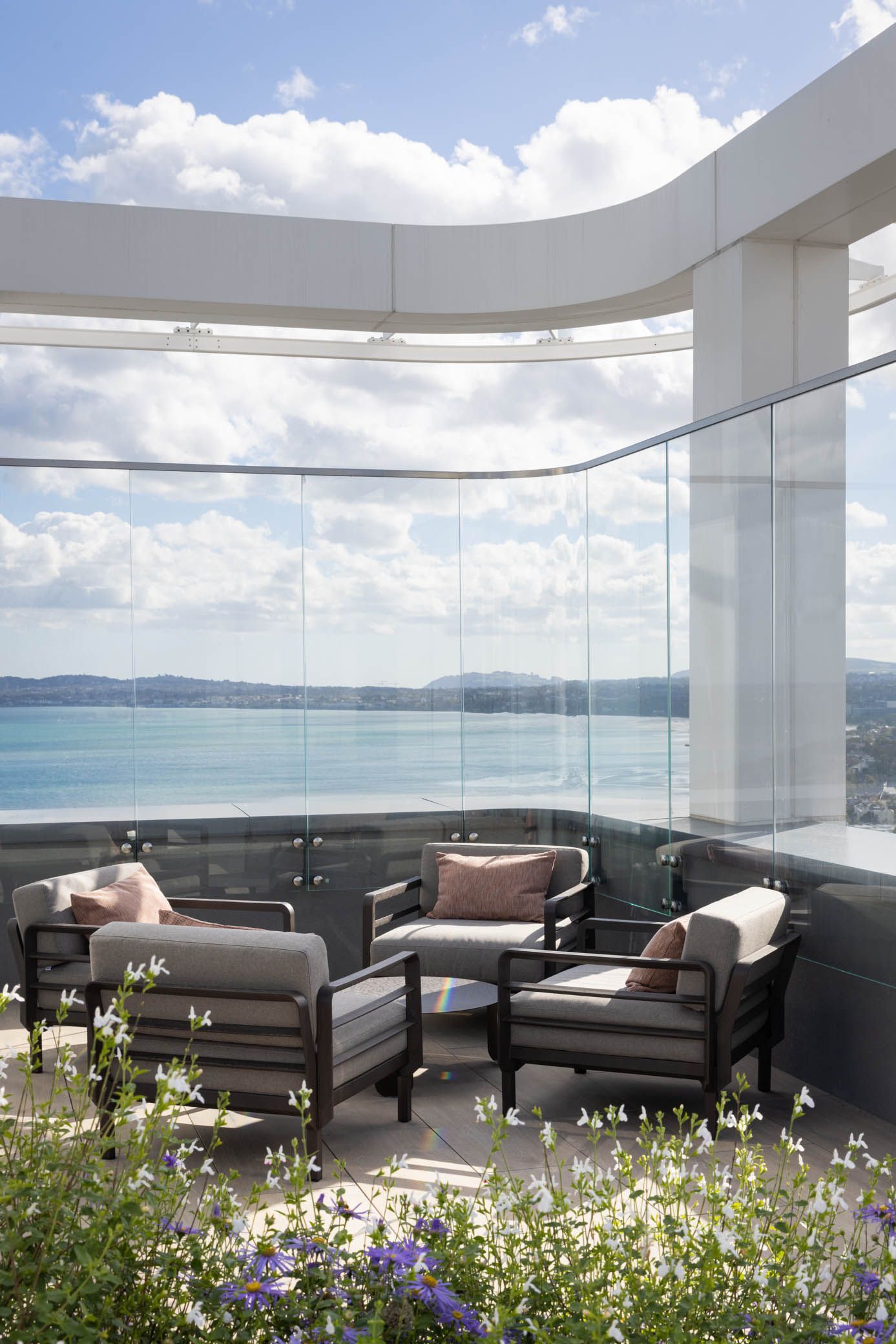 Patio lounge with cushioned chairs overlooking a calm ocean and cloudy sky, framed by glass walls and flowers
