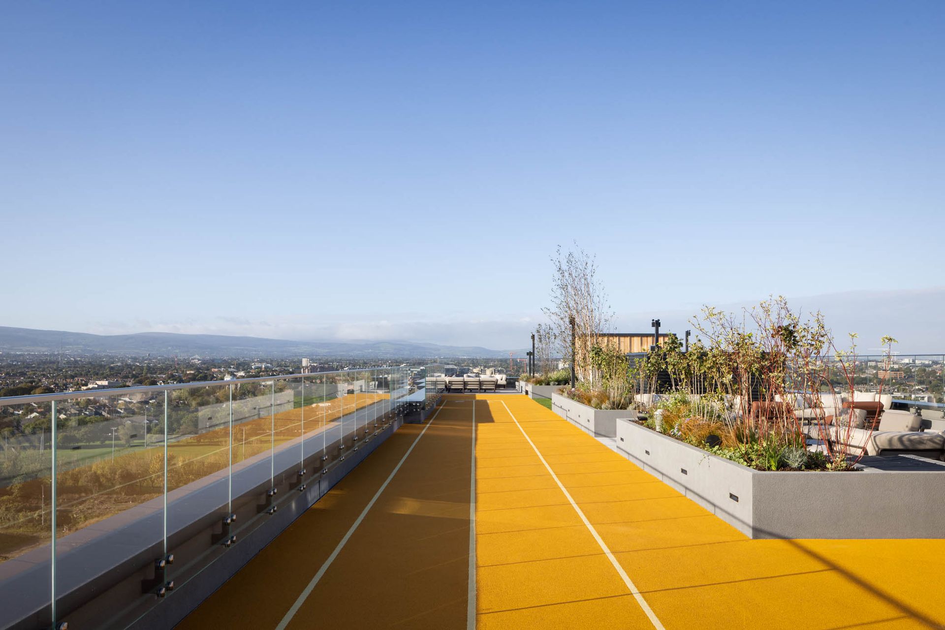 Rooftop terrace with bright yellow walkway, city skyline in the distance, and glass railing under a clear blue sky