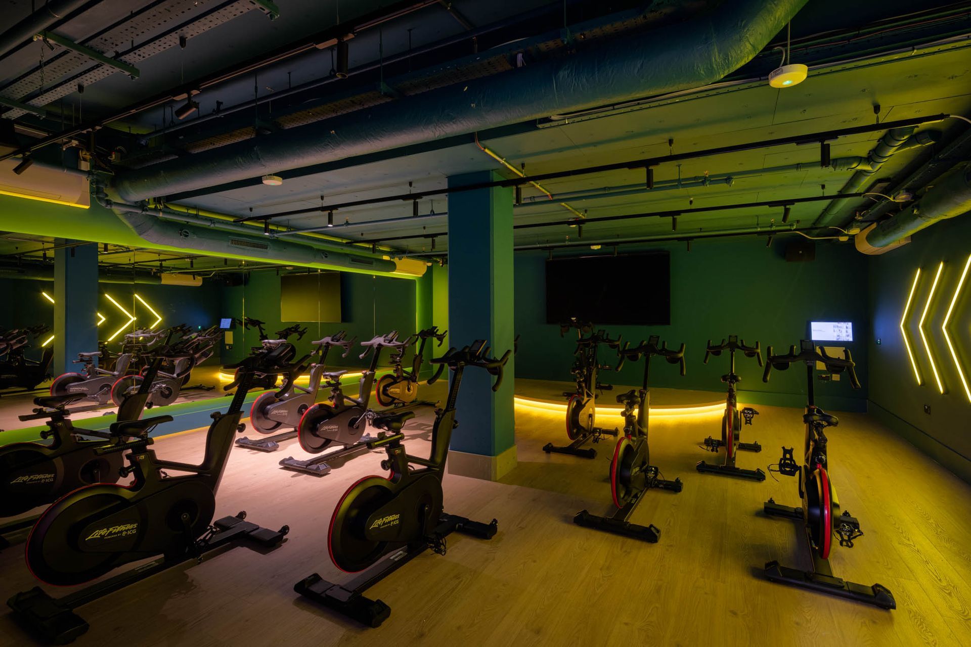 Indoor cycling studio with stationary bikes, neon green lighting, and exposed ceiling pipes