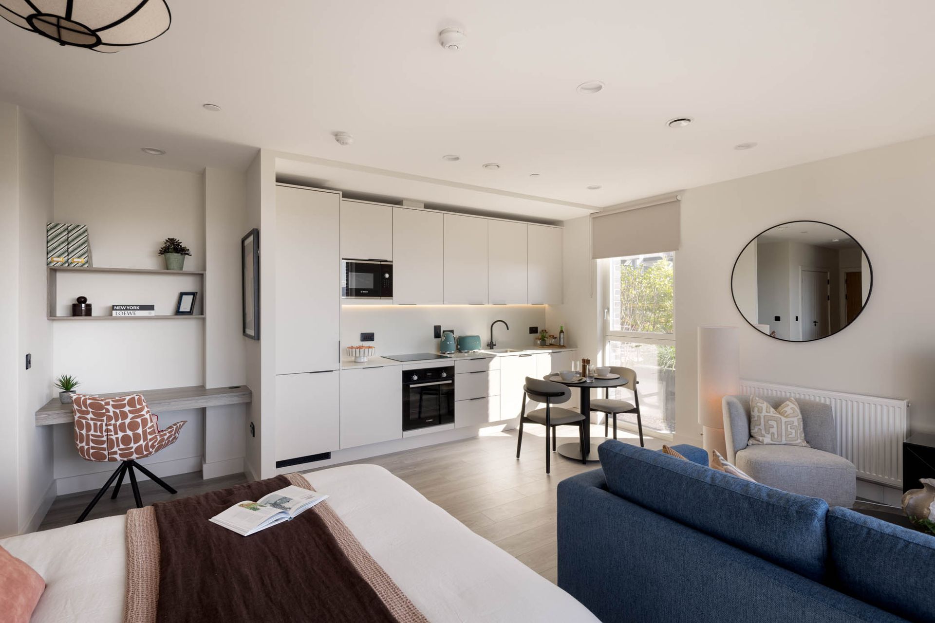 Modern open-plan apartment with white kitchen, blue sofa, dining table, and wall mirror