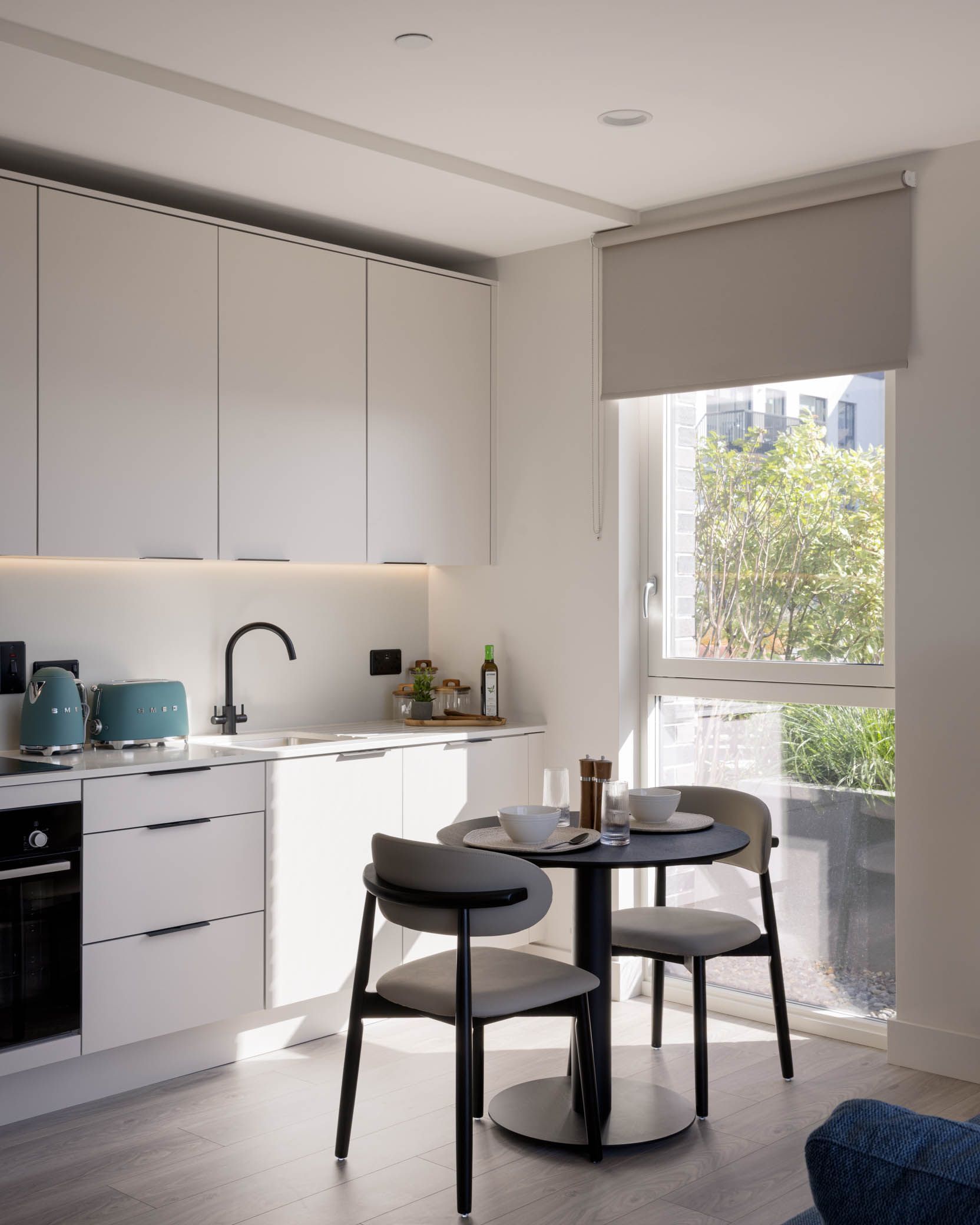 Modern white kitchen with round table, black chairs, and a large window overlooking greenery