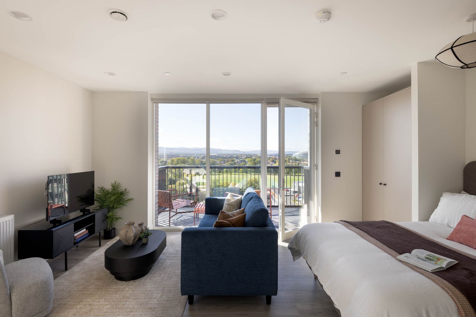 Bright modern living room with sofa, TV, bed, and balcony doors overlooking a city view