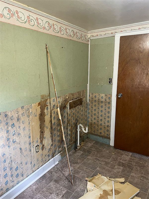 A room with peeling wallpaper, exposed pipes, and a door; walls are green and patterned.