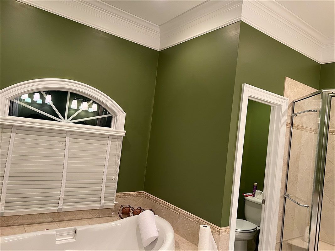 Green-walled bathroom with a bathtub, arched window with shutters, and a toilet visible through the doorway.