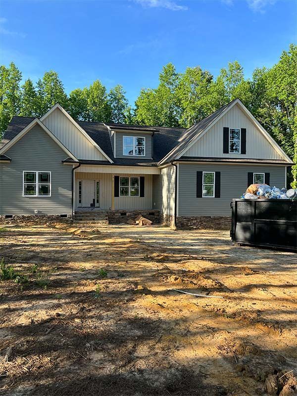 New construction house, gray siding, white trim, with black shutters, on a lot with dirt and trees.