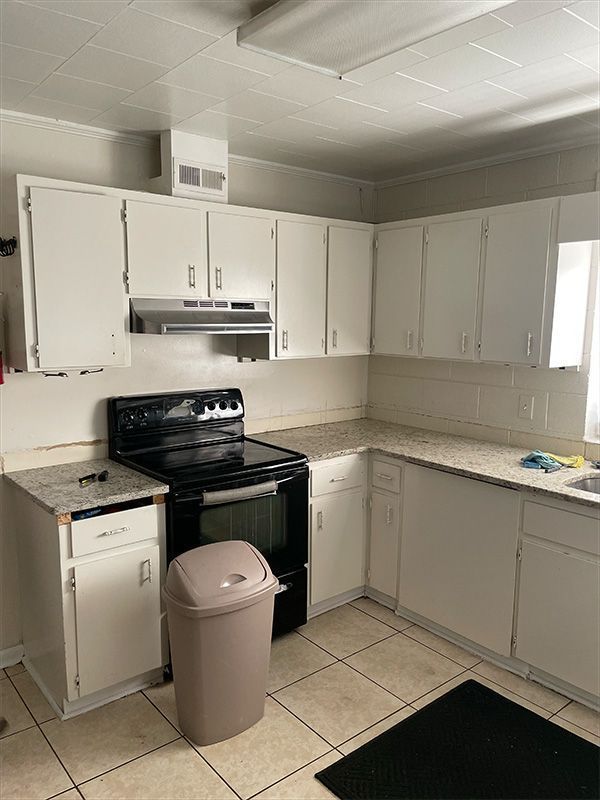 Small white kitchen with black stove, white cabinets, beige tile floor, and trash can in foreground
