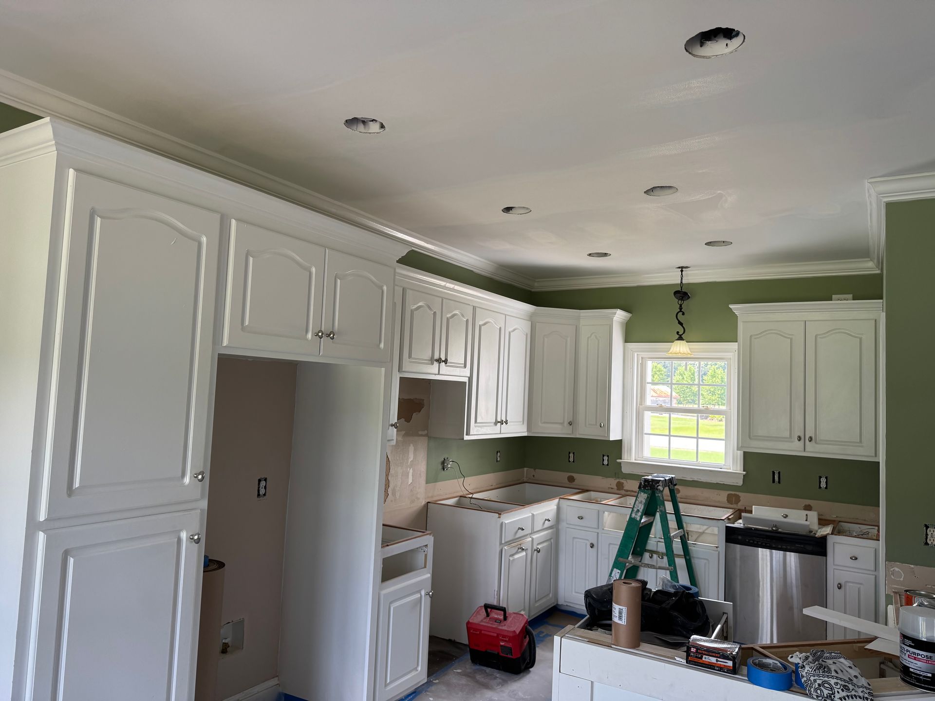 Kitchen under renovation with white cabinets, green walls, a ladder, and tools on the floor.