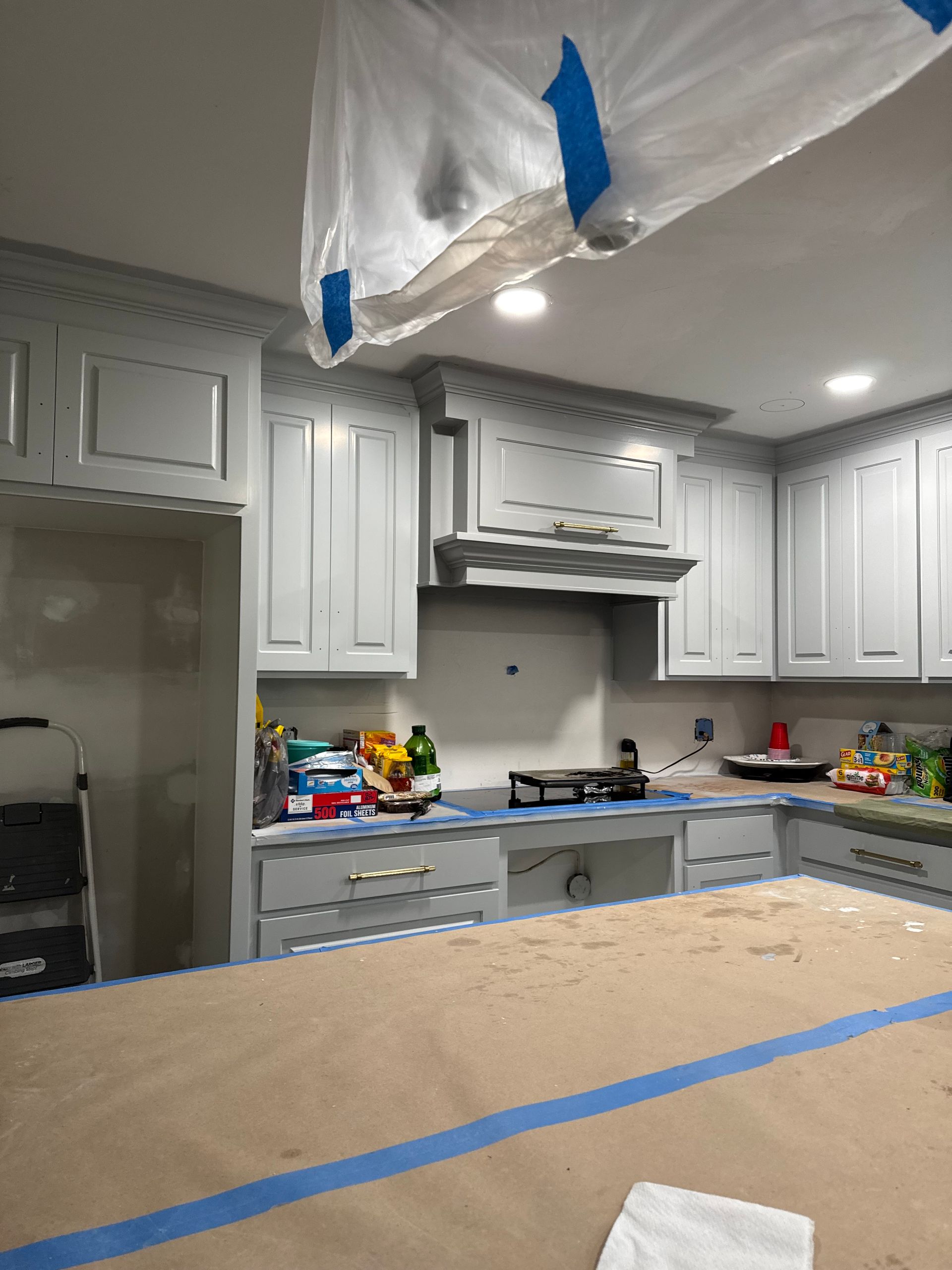 Kitchen under renovation with white cabinets, covered appliances, and blue painter’s tape on countertops and ceiling plastic