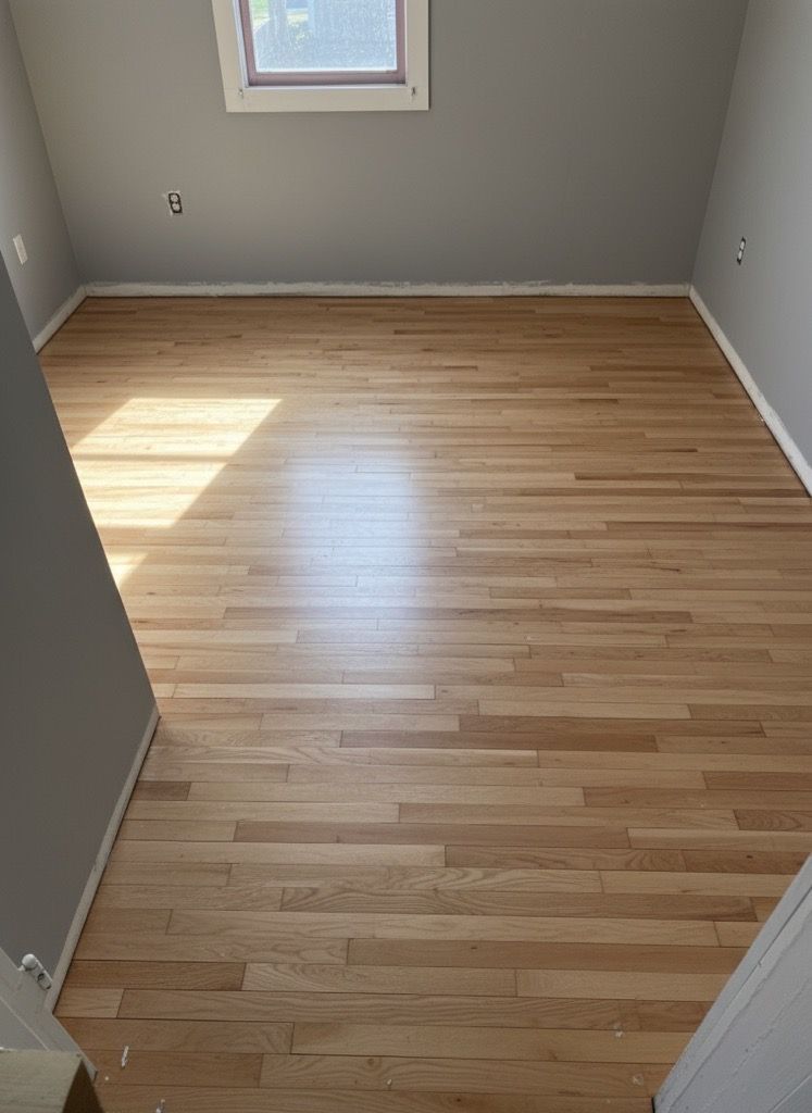 Empty room with light wood floor, gray walls, and a small window letting in sunlight.