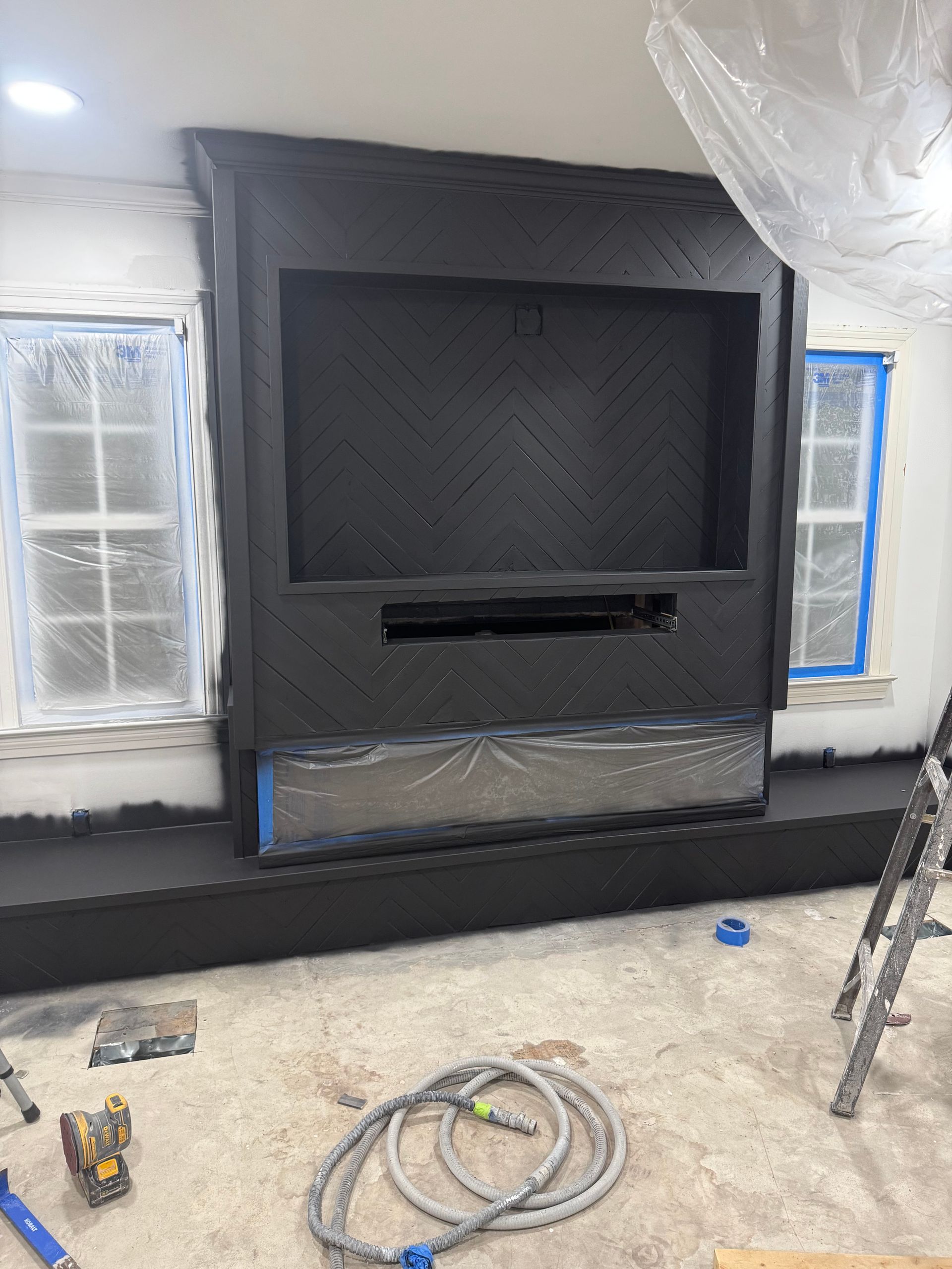 Construction site with a black wall-mounted fireplace surround framed by protective plastic and tools on the floor.