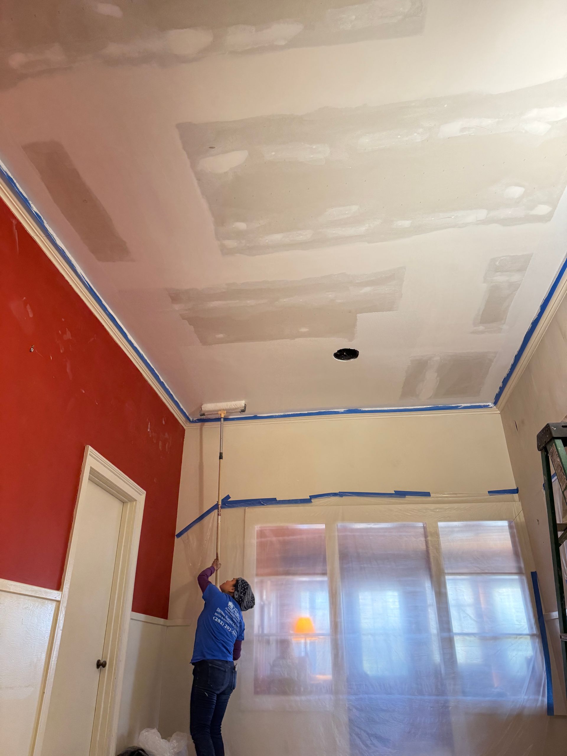 Person painting or repairing a room ceiling and walls, with taped edges and a covered window.
