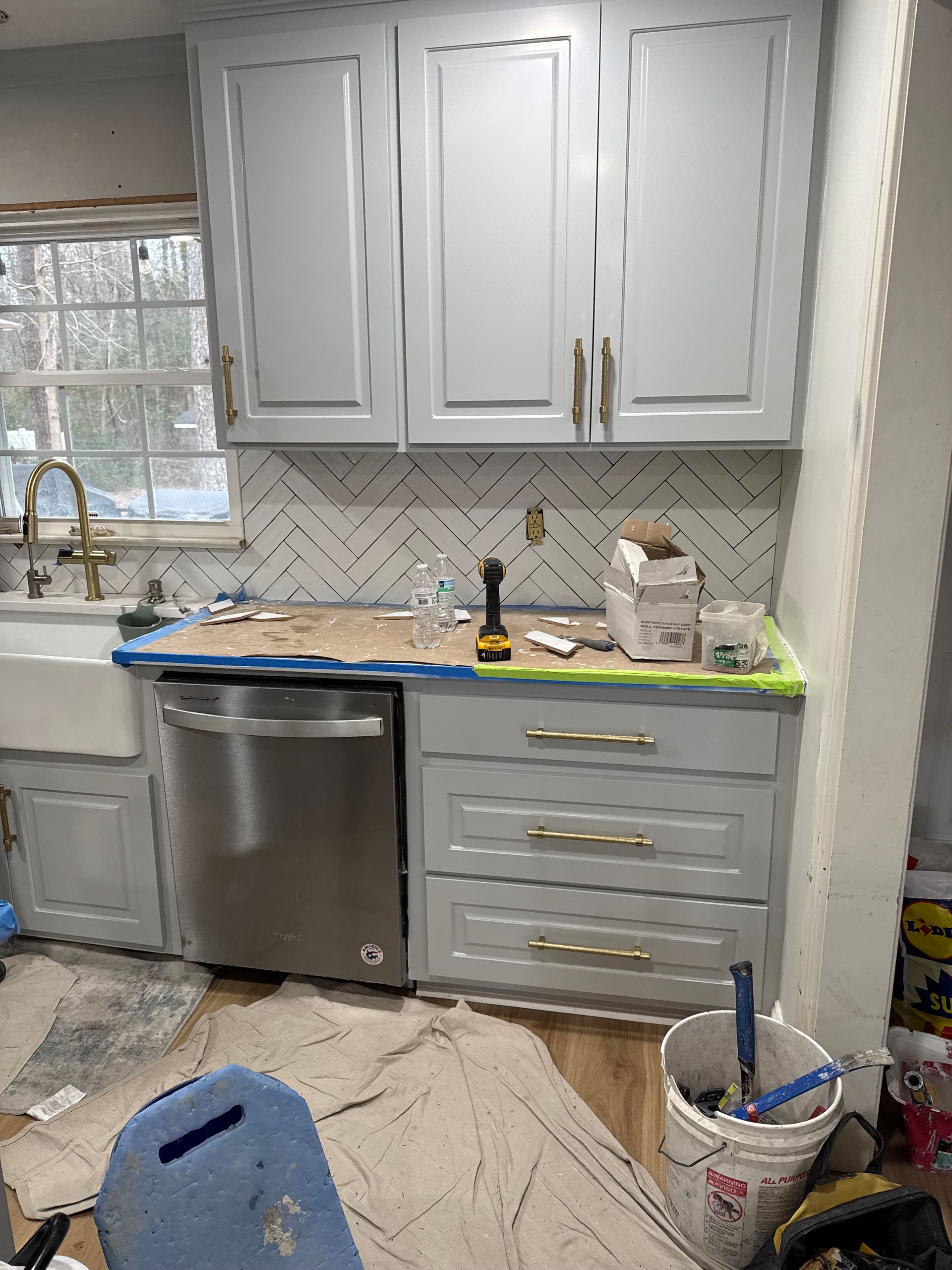 Small kitchen under renovation with white cabinets, stainless dishwasher, blue bucket, and cleaning supplies on floor