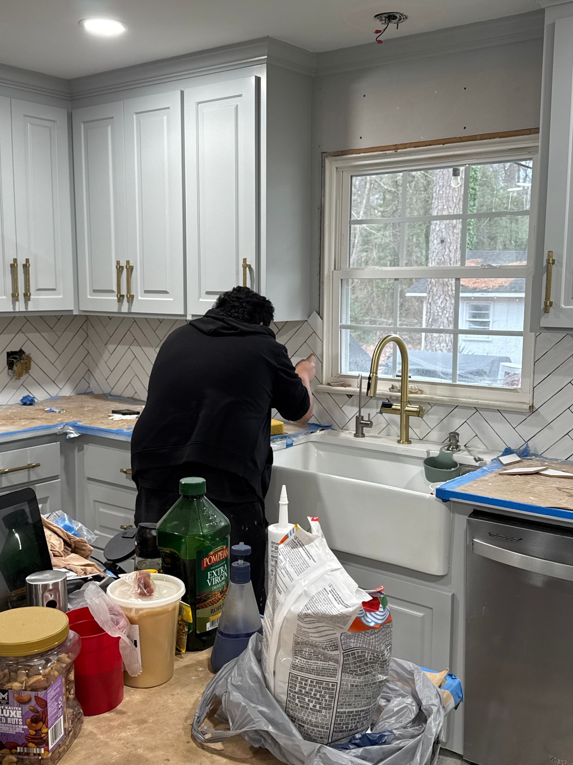 Person at kitchen sink, leaning over counter under a window in a white kitchen.