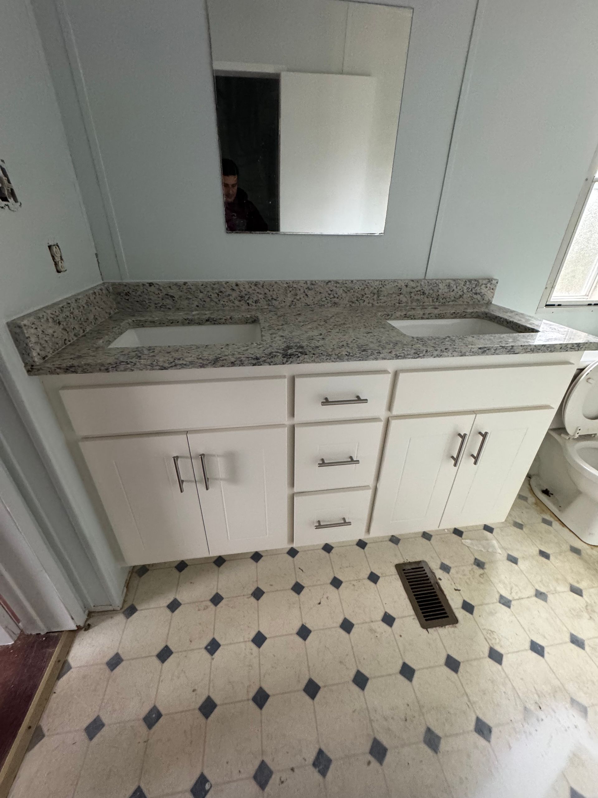 White bathroom vanity with granite countertop, mirror, and floor vent on patterned tile floor