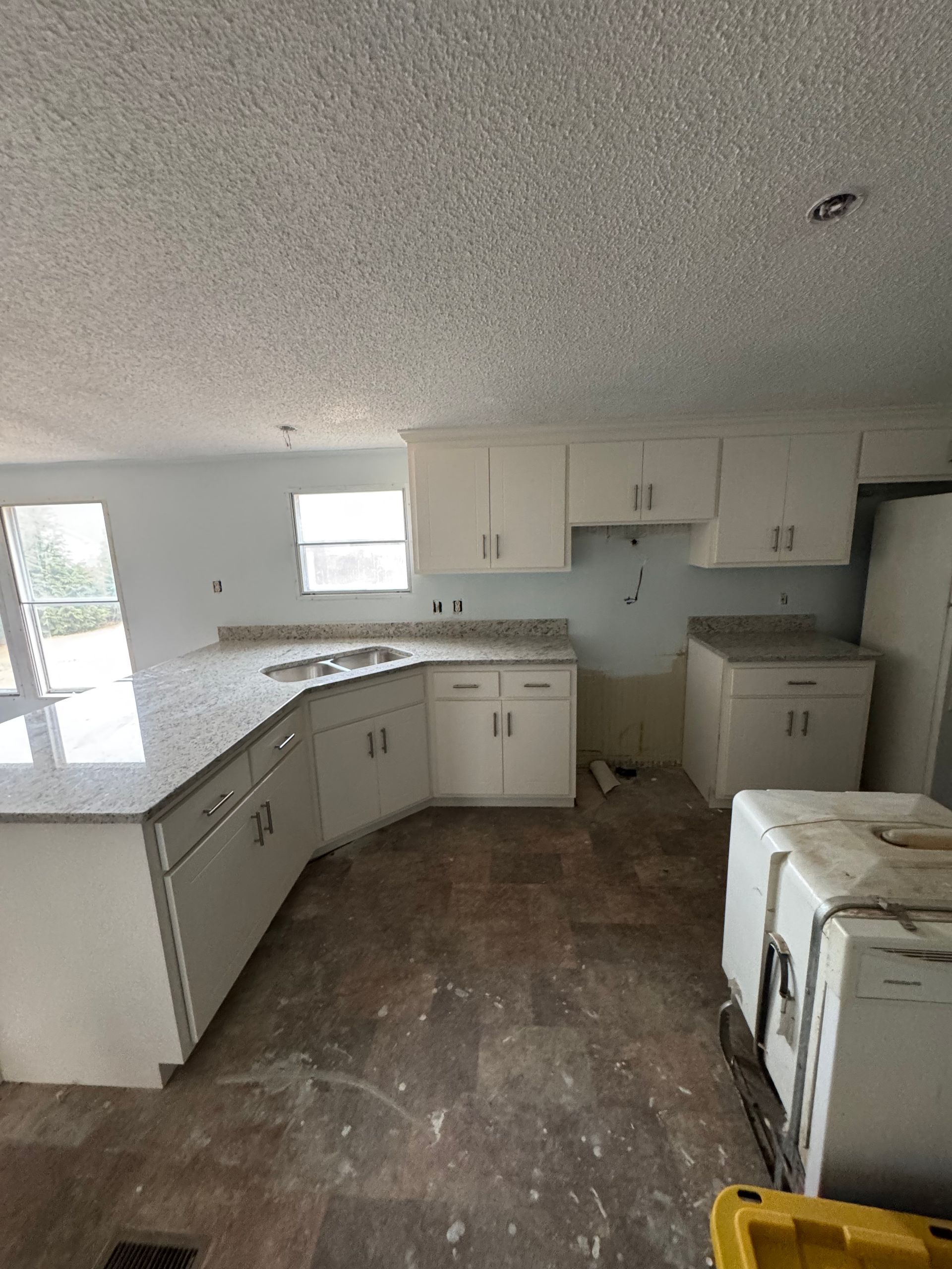 Empty kitchen with white cabinets, exposed flooring, and a window by the sink.