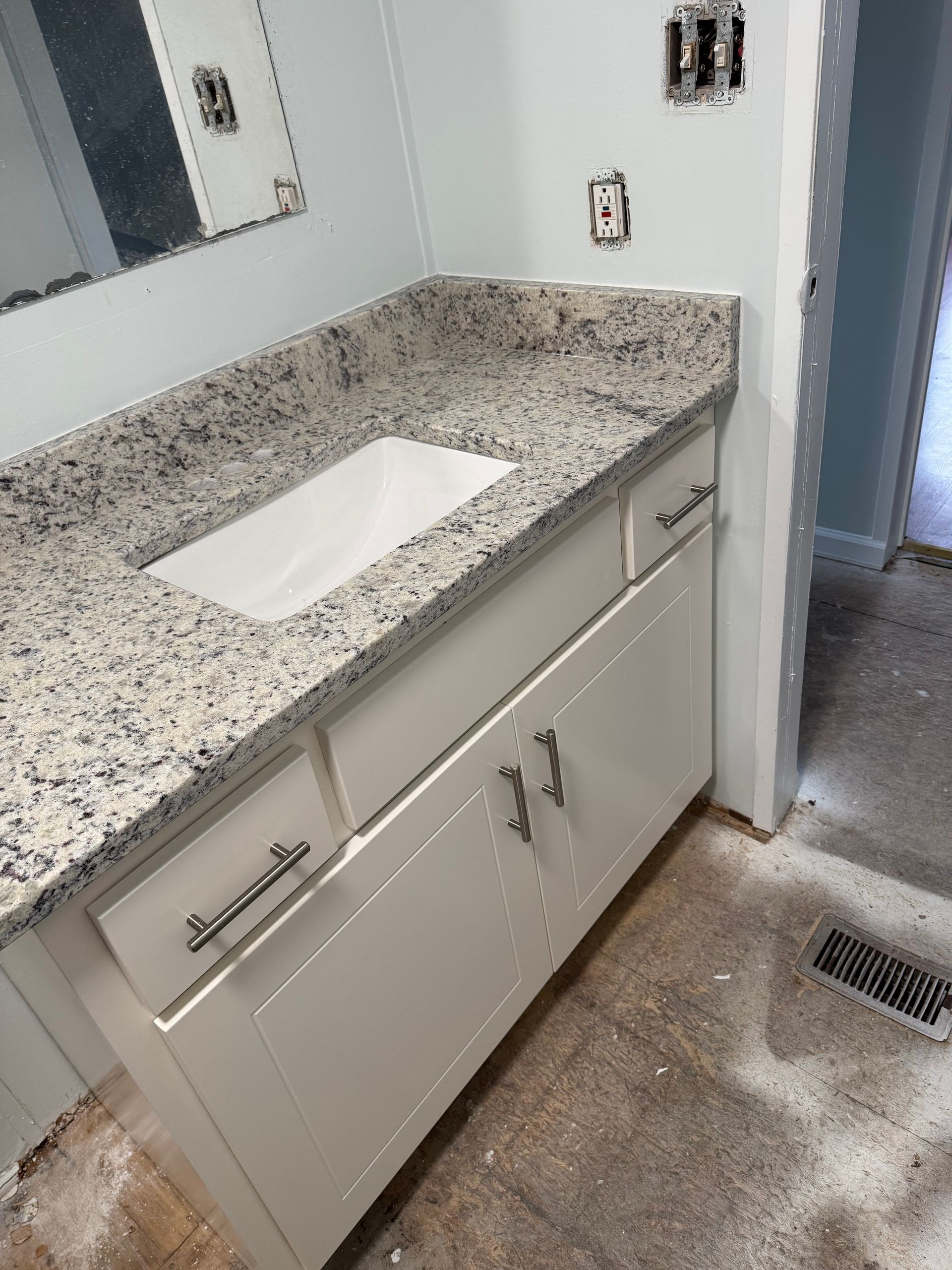 Bathroom vanity with granite countertop, sink cutout, and white cabinets beside a floor vent.