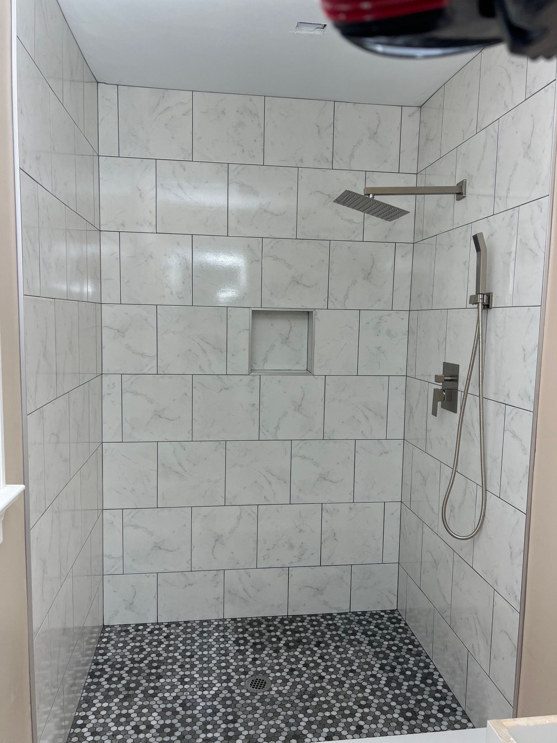 White tiled shower with black patterned floor, rainfall showerhead, and wall-mounted handheld sprayer.