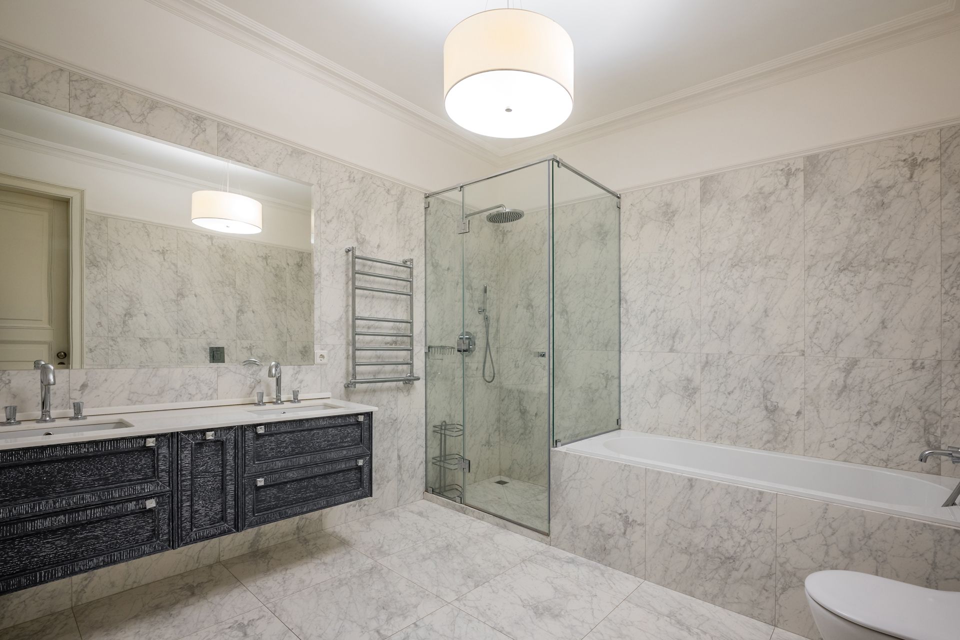 Modern bathroom with marble walls, a glass shower, a bathtub, and a dark vanity with double sinks.