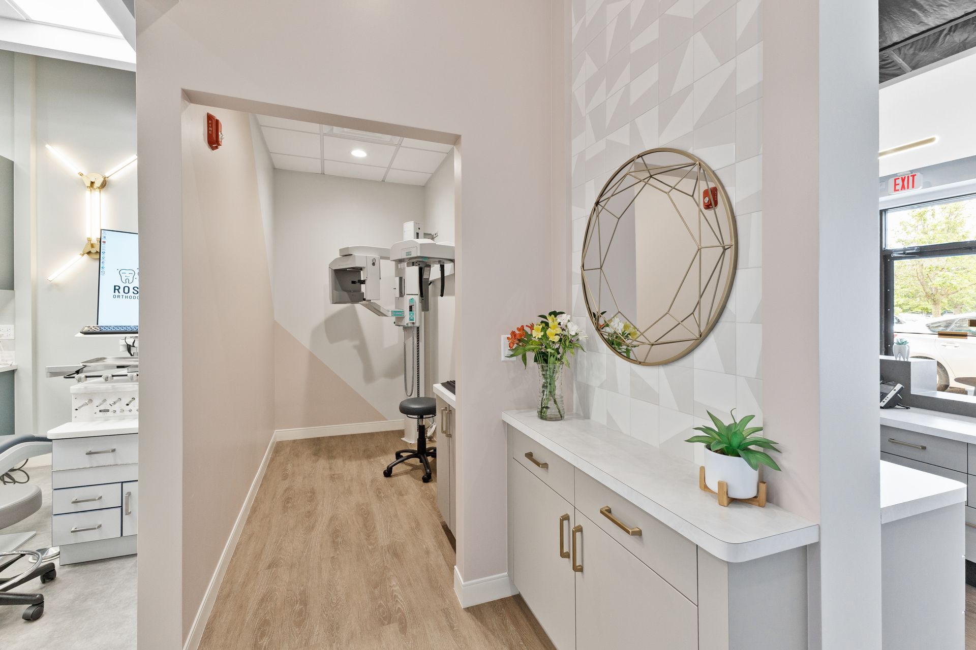 Bright clinic hallway with a reception cabinet, wall mirror, and treatment room in the background