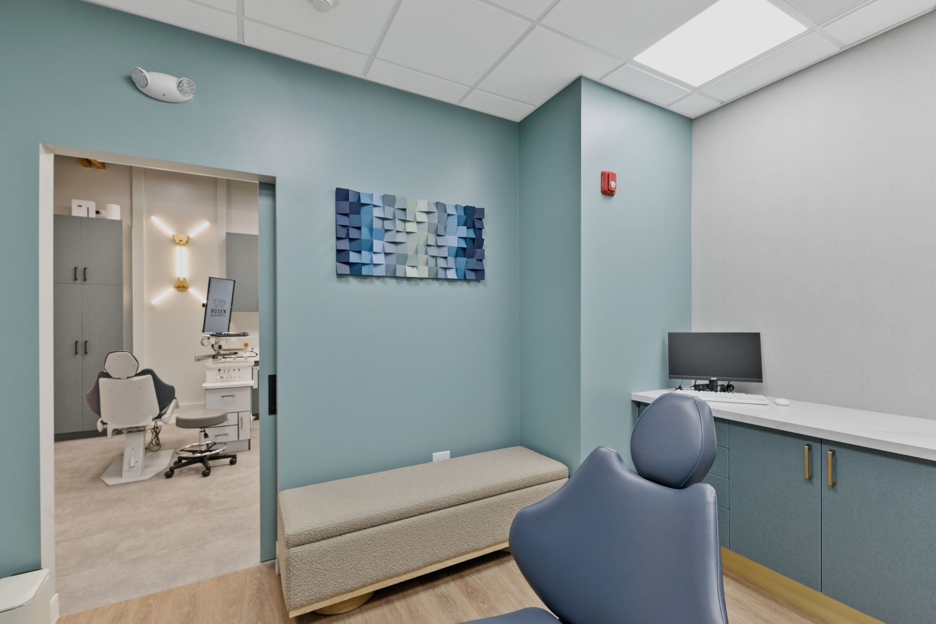 Modern medical exam room with teal walls, exam chair, desk, and open doorway to dental equipment