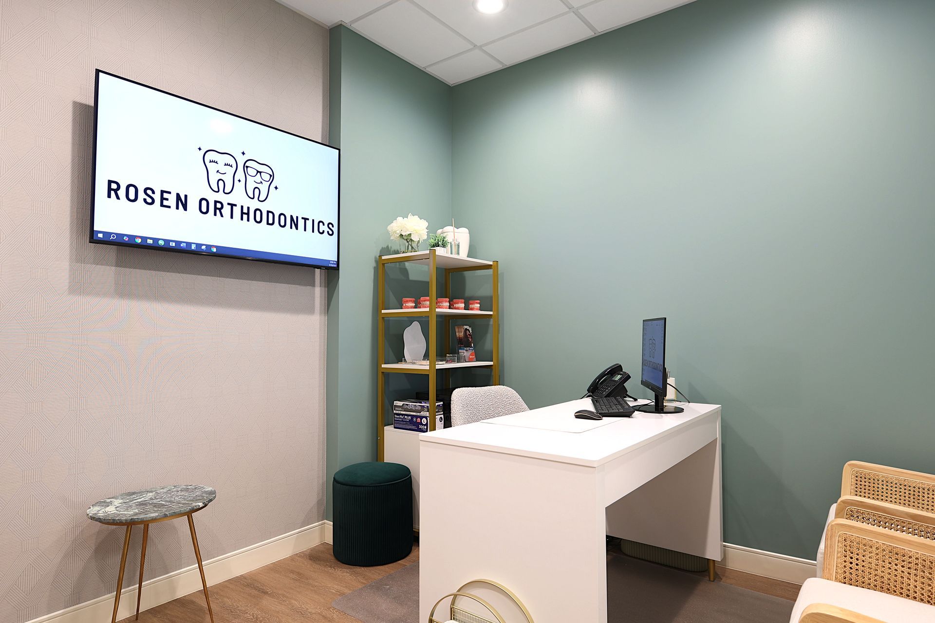 Small dental or clinic reception area with a desk, screen, shelves, and a wall sign reading “Roson Orthodontics.”