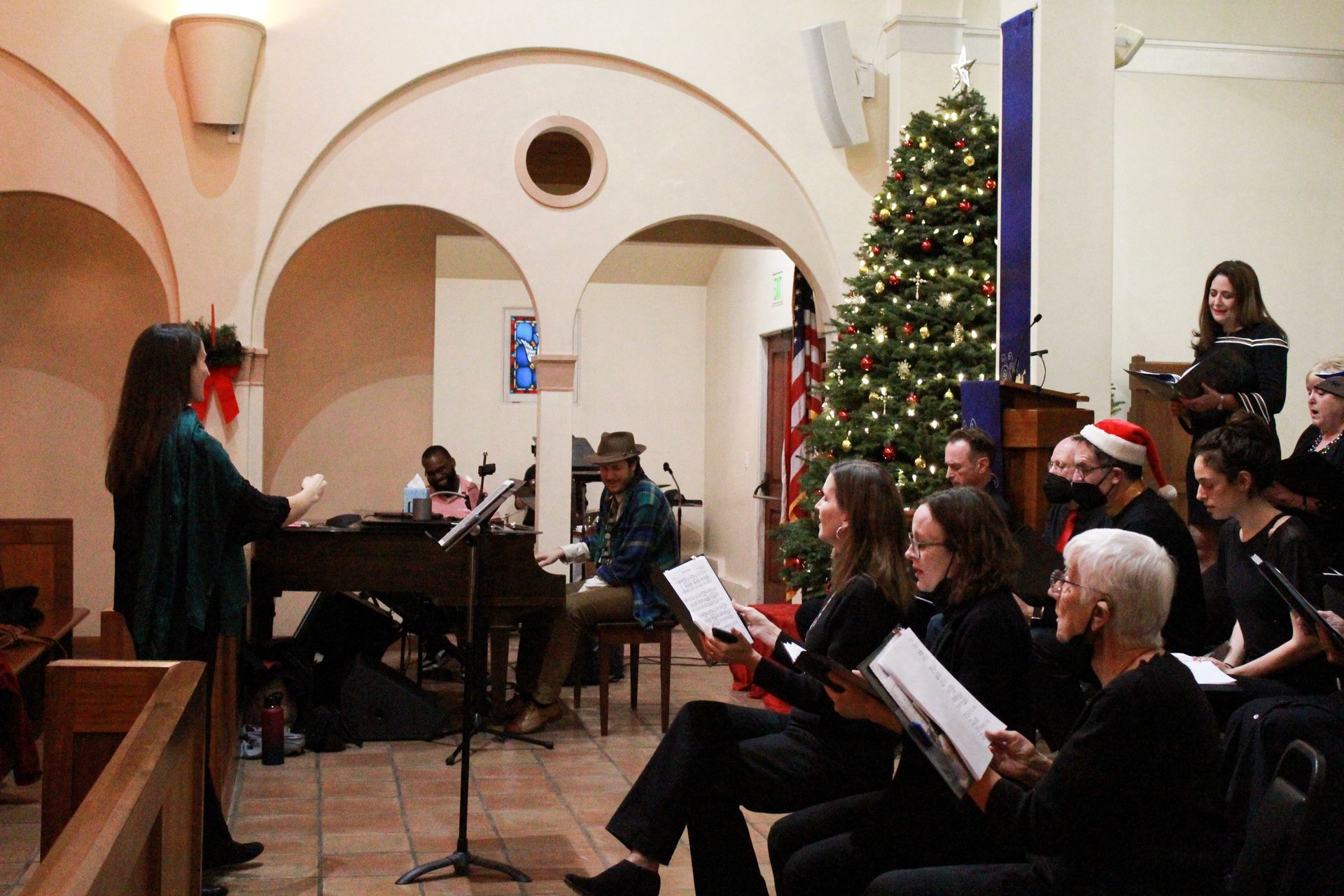 Choir singing in a church with a Christmas tree. Conductor leads. People sit and stand, holding sheet music.