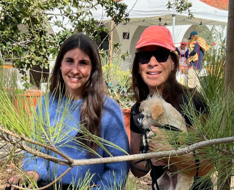 Two smiling women, one holding a small dog, stand near greenery outdoors with a white tent in the background.