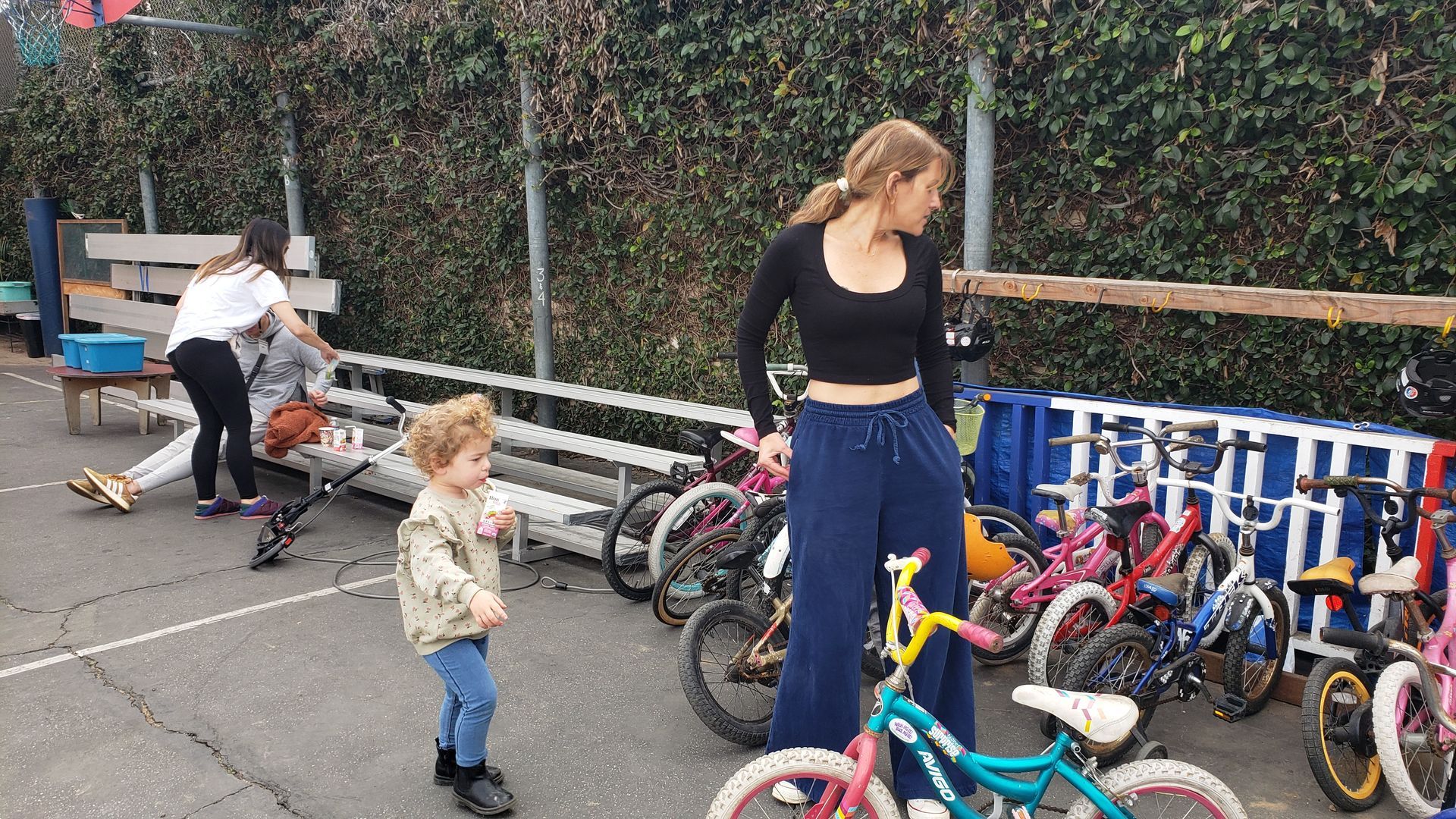 A child and woman near bicycles outdoors; others in background. The child dances, the woman looks.