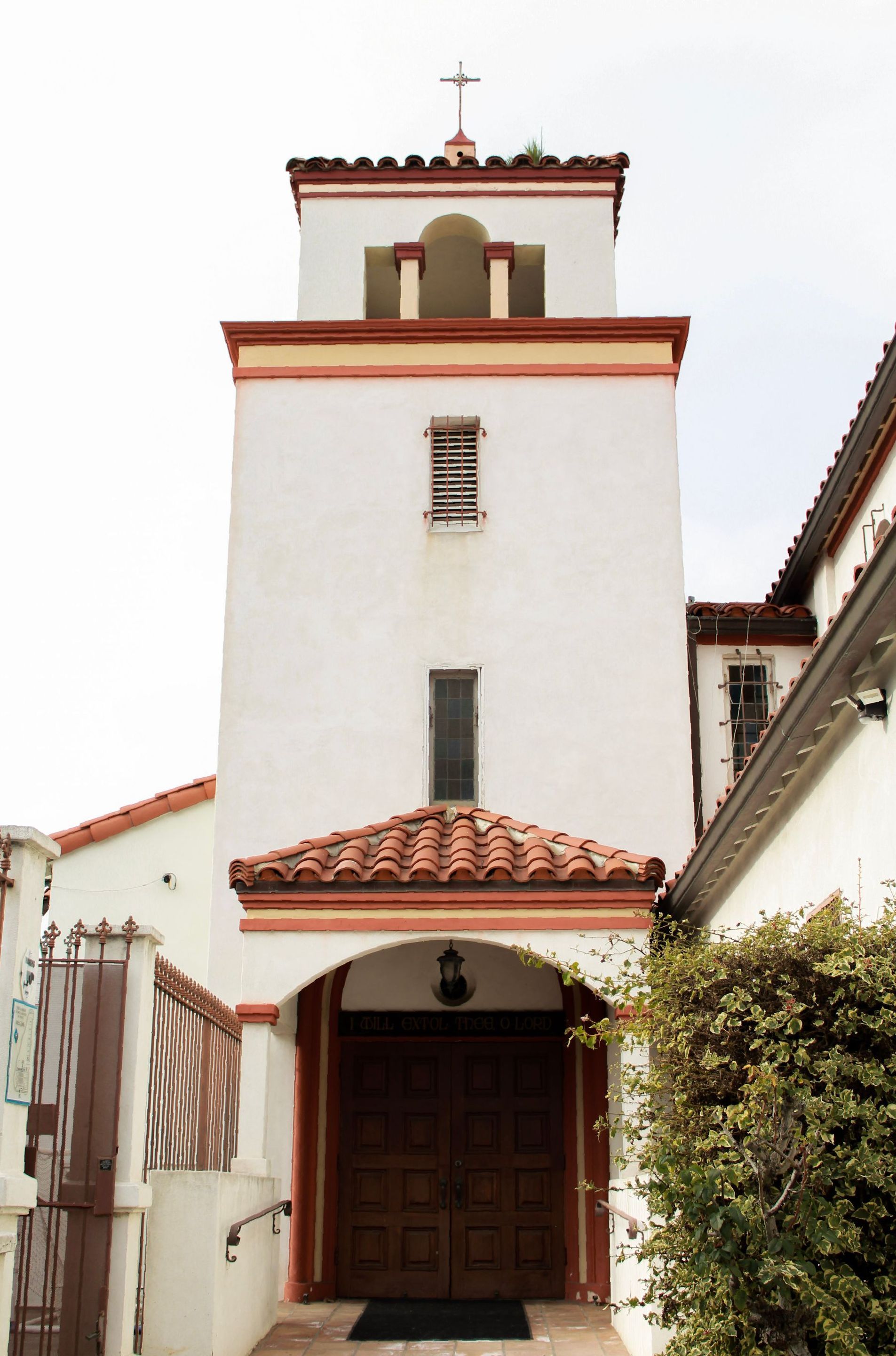 White stucco church tower with terracotta roof and trim, arched entrance, cross at top.